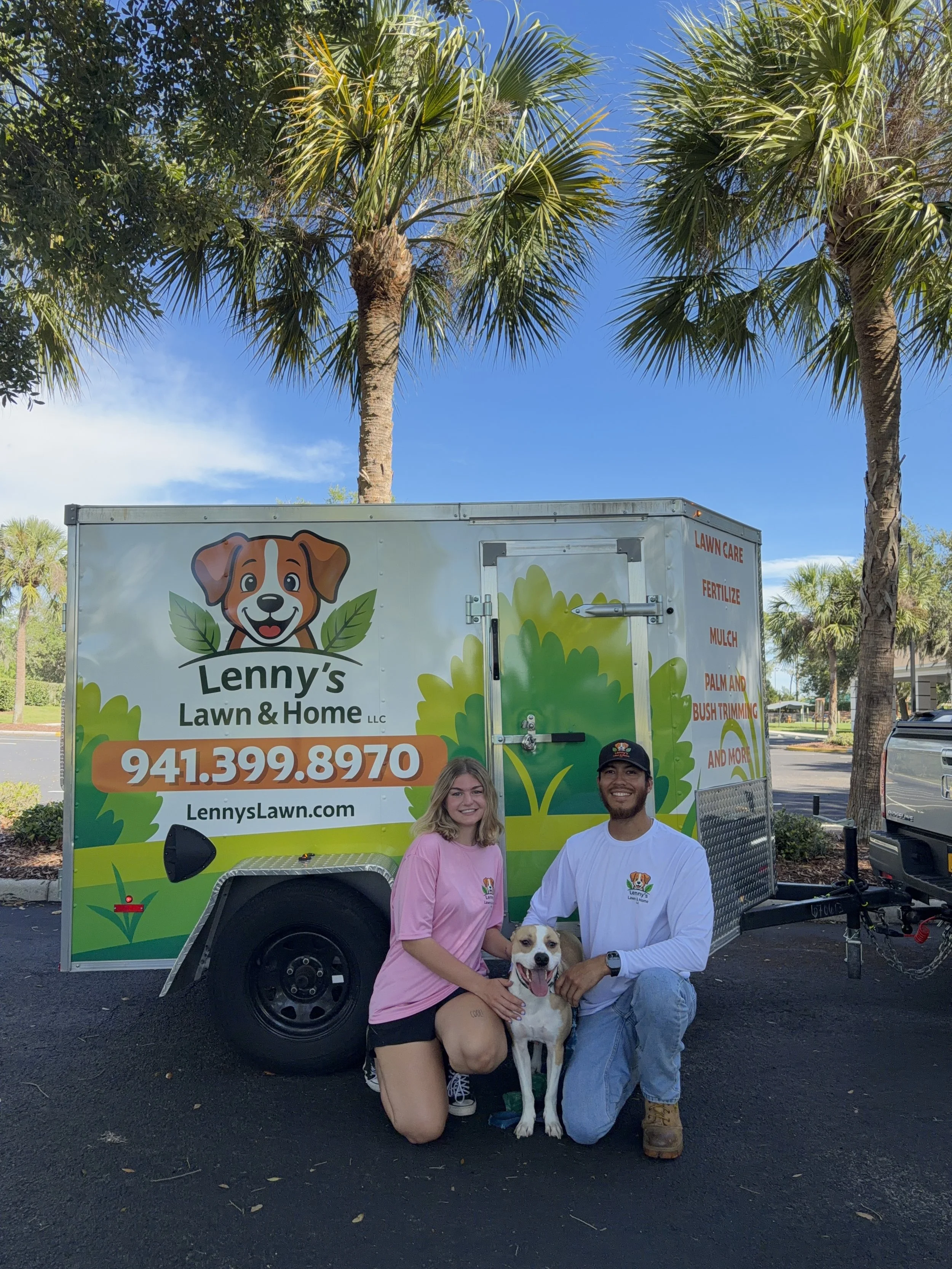 Two people kneeling with a dog in front of a lawn care trailer, with palm trees and a blue sky in the background.