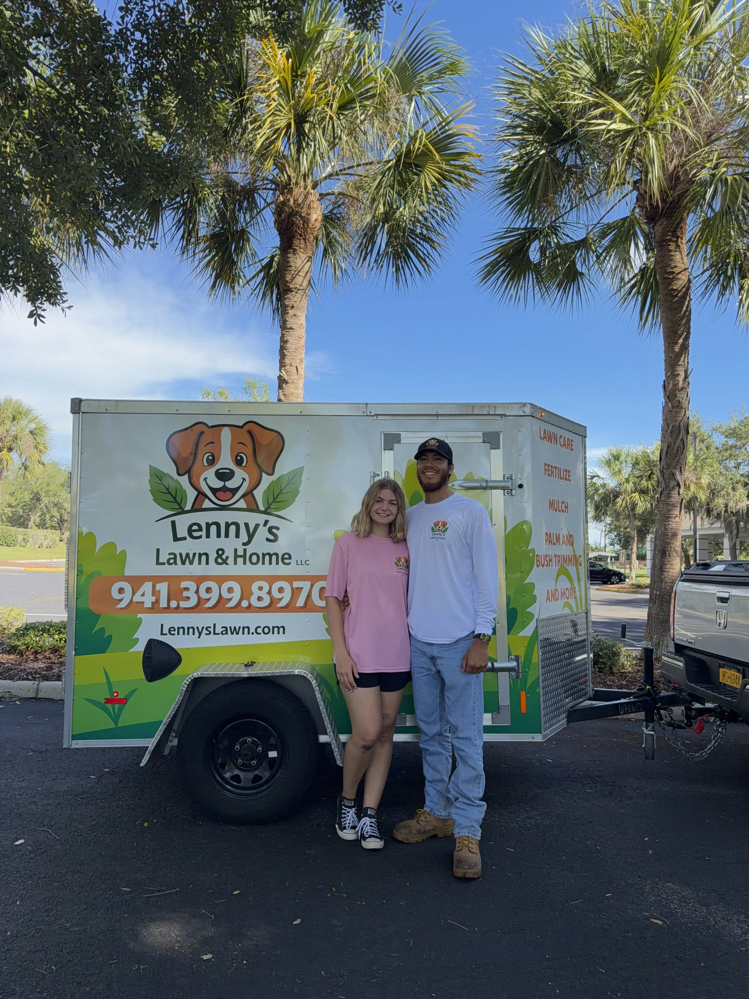 A man and a woman stand together in front of a colorful mobile for Lenny's Lawn & Home, surrounded by palm trees and a clear blue sky. The man is wearing a white long sleeve shirt, light blue jeans, tan work boots, and a black cap. The woman is wearing a pink sweatshirt, black shorts, and black Converse sneakers.