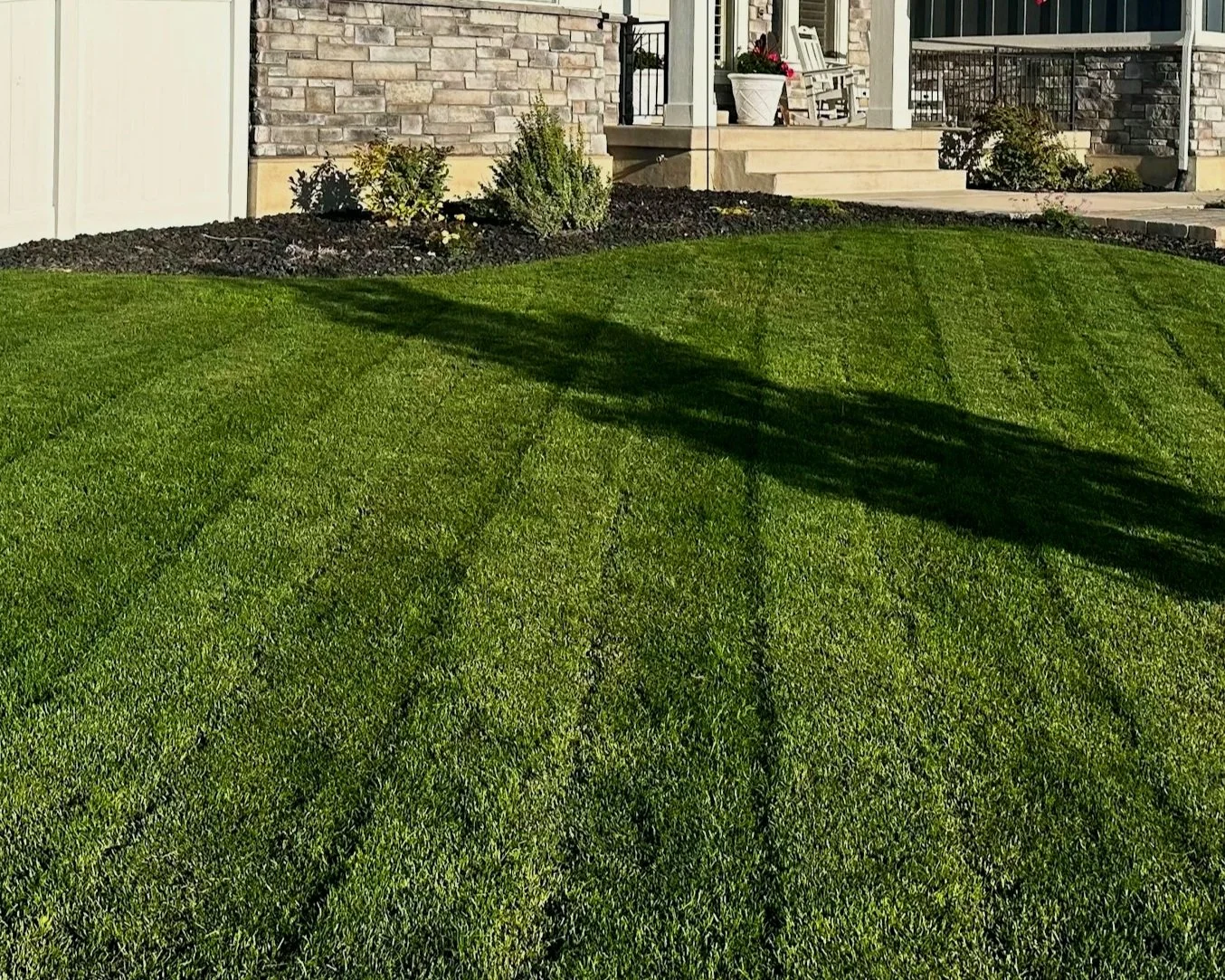 Well-maintained green lawn with freshly mowed grass and visible mowing lines, in front of a house with a small porch, stairs, and flower pots, surrounded by flower beds with shrubs and mulch.