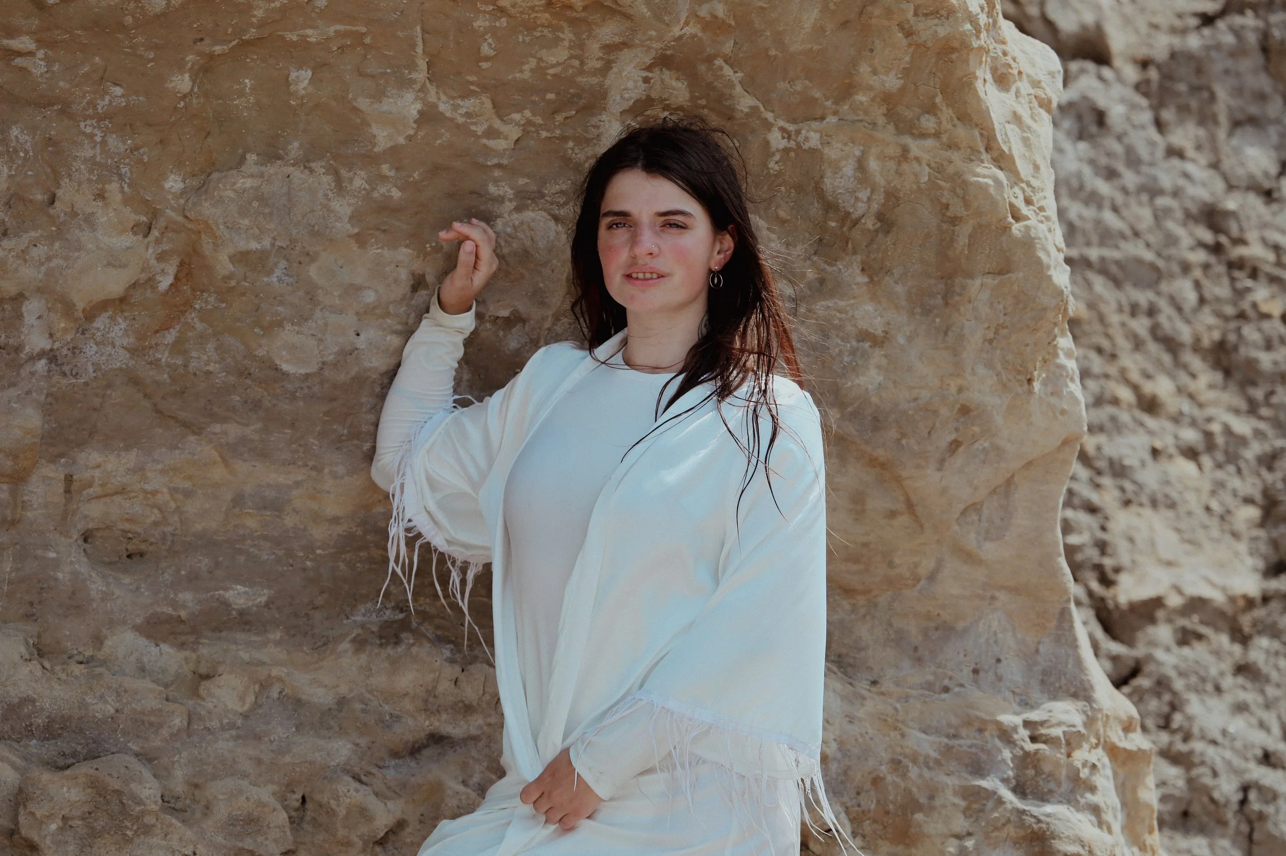 Femme avec des cheveux bruns, porter un vêtement blanc, posant près d'une falaise rocheuse aride.