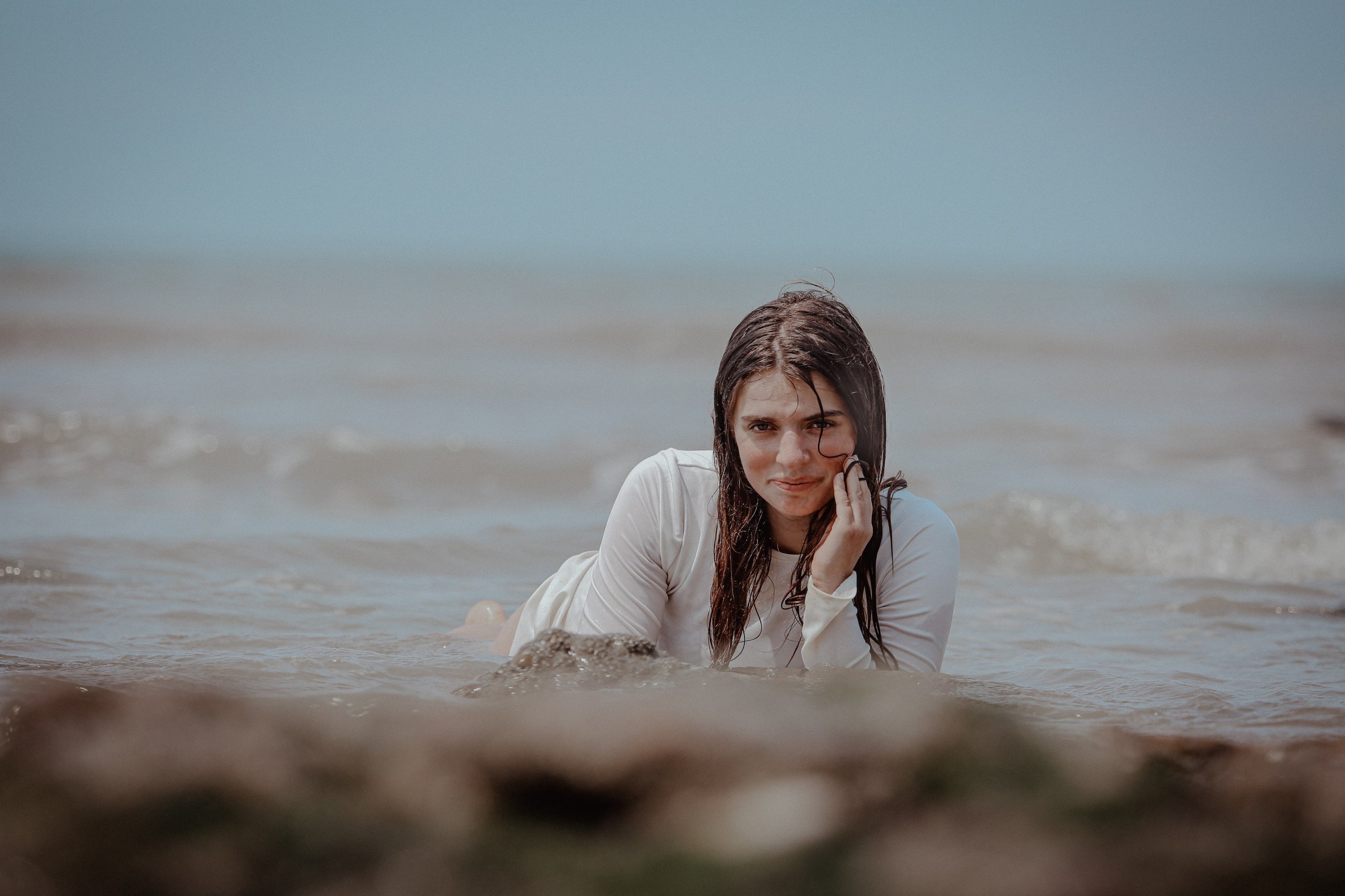 Jeune femme à la plage, allongée dans l'eau, portant une chemise blanche, avec la mer et le ciel en arrière-plan.