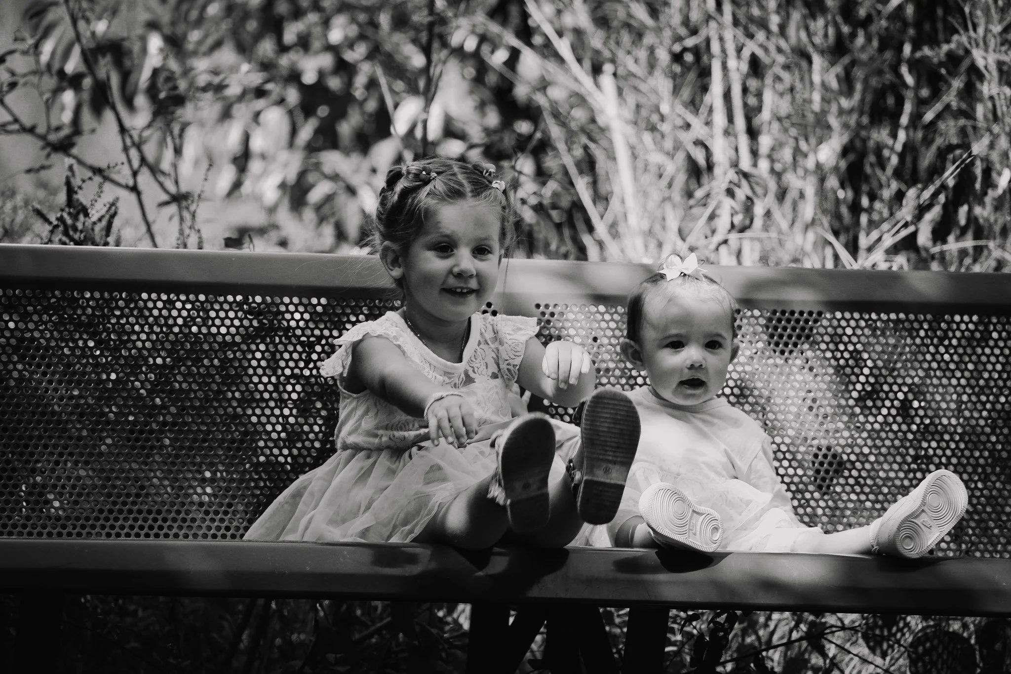 Deux jeunes filles assises sur un banc en métal dans un environnement naturel, l'une porte une robe claire avec des détails en dentelle, l'autre porte une robe claire avec un gros ruban dans les cheveux. Elles sourient et regardent vers l'appareil ph