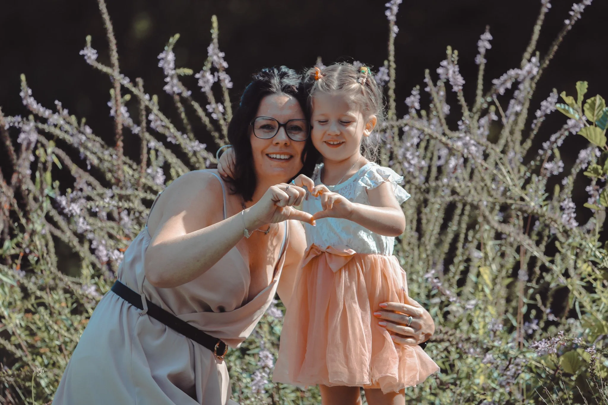 Une femme et une jeune fille forment un cœur avec leurs mains devant un arrière-plan de fleurs violettes.