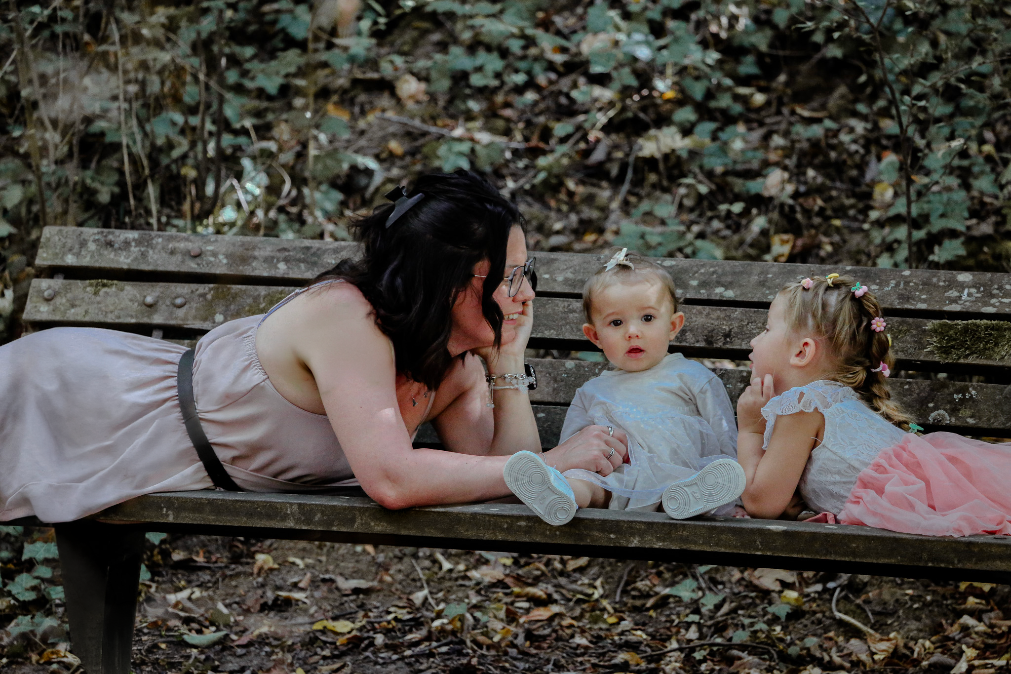 Une femme avec deux jeunes filles assises sur un banc en bois dans la forêt. La femme parle à la plus jeune, tandis que la plus âgée écoute. Les filles portent des robes de fête avec des fleurs dans leurs cheveux.