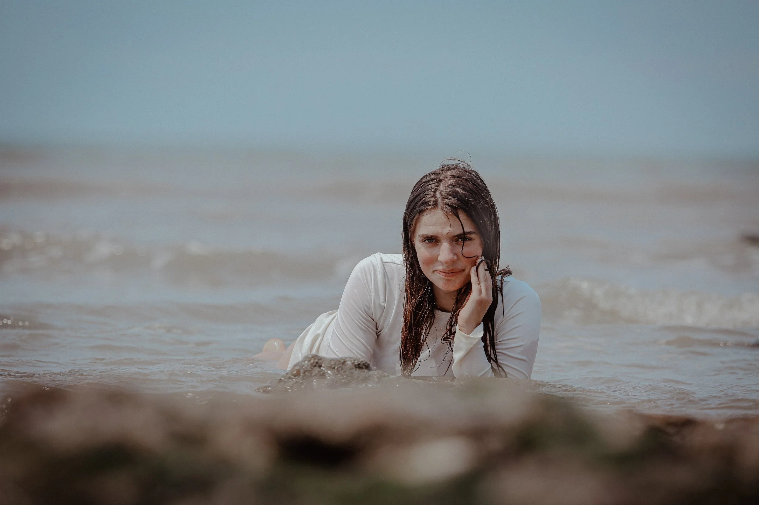 Jeune femme à la plage, allongée dans l'eau, cheveux mouillés, regardant vers l'avant, tenue blanche, avec le ciel bleu en arrière-plan.