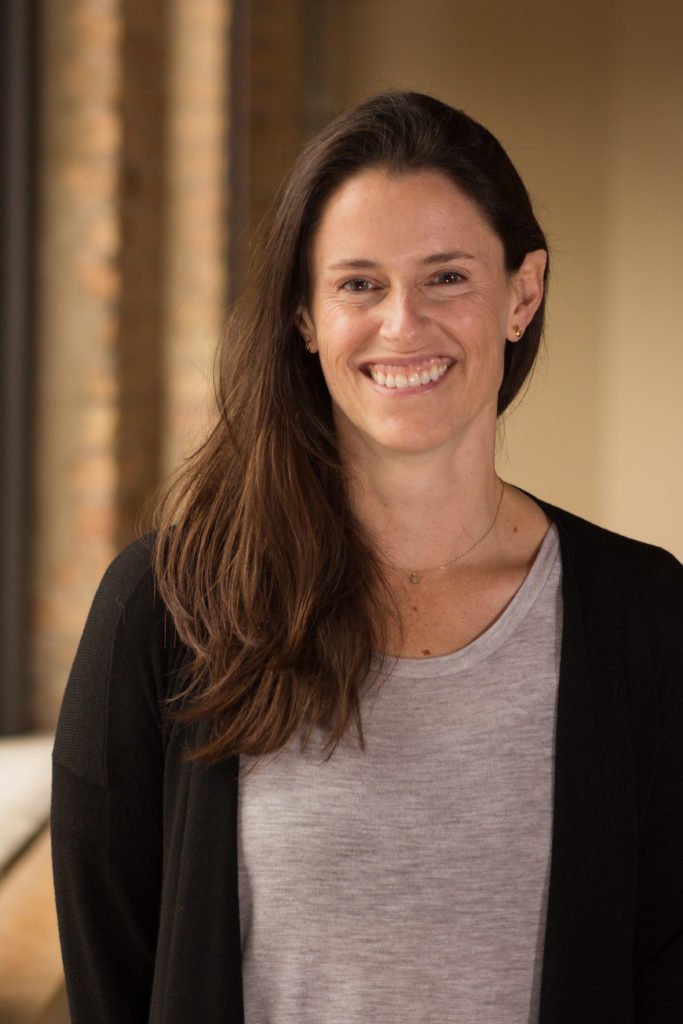 A woman with long brown hair and fair skin, smiling at the camera, wearing a light gray shirt and a black cardigan, standing indoors with a blurred background.