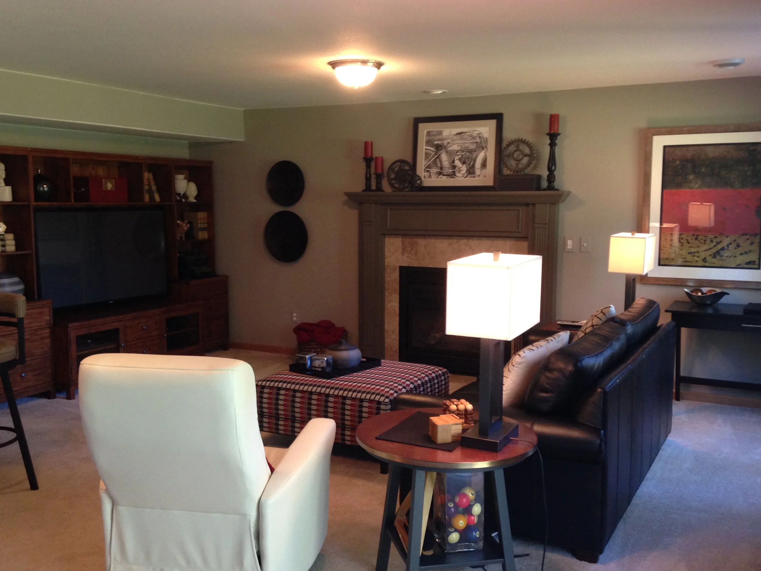 A living room with a white armchair, a round wooden table with wood blocks, a sofa with patterned pillows, a black side table with colorful balls in a glass container, a fireplace with a mantle decorated with framed art, candles, and gear decor, a large abstract painting on the wall, a wooden bookshelf holding books and decor, and a ceiling light fixture.