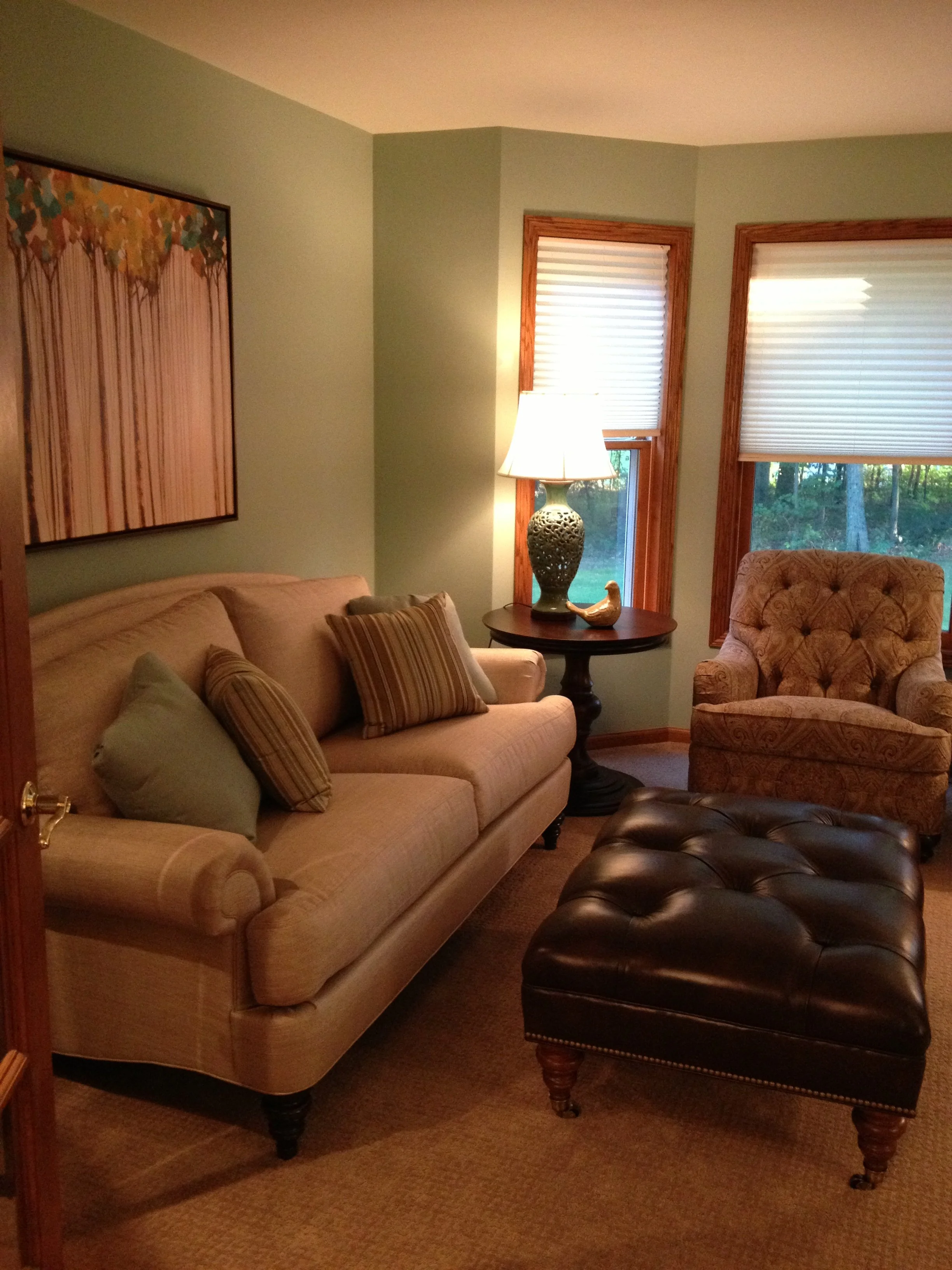 Living room with beige sofa, striped pillows, patterned armchair, leather ottoman, table with lamp and decorative duck, and windows with wooden frames and blinds.