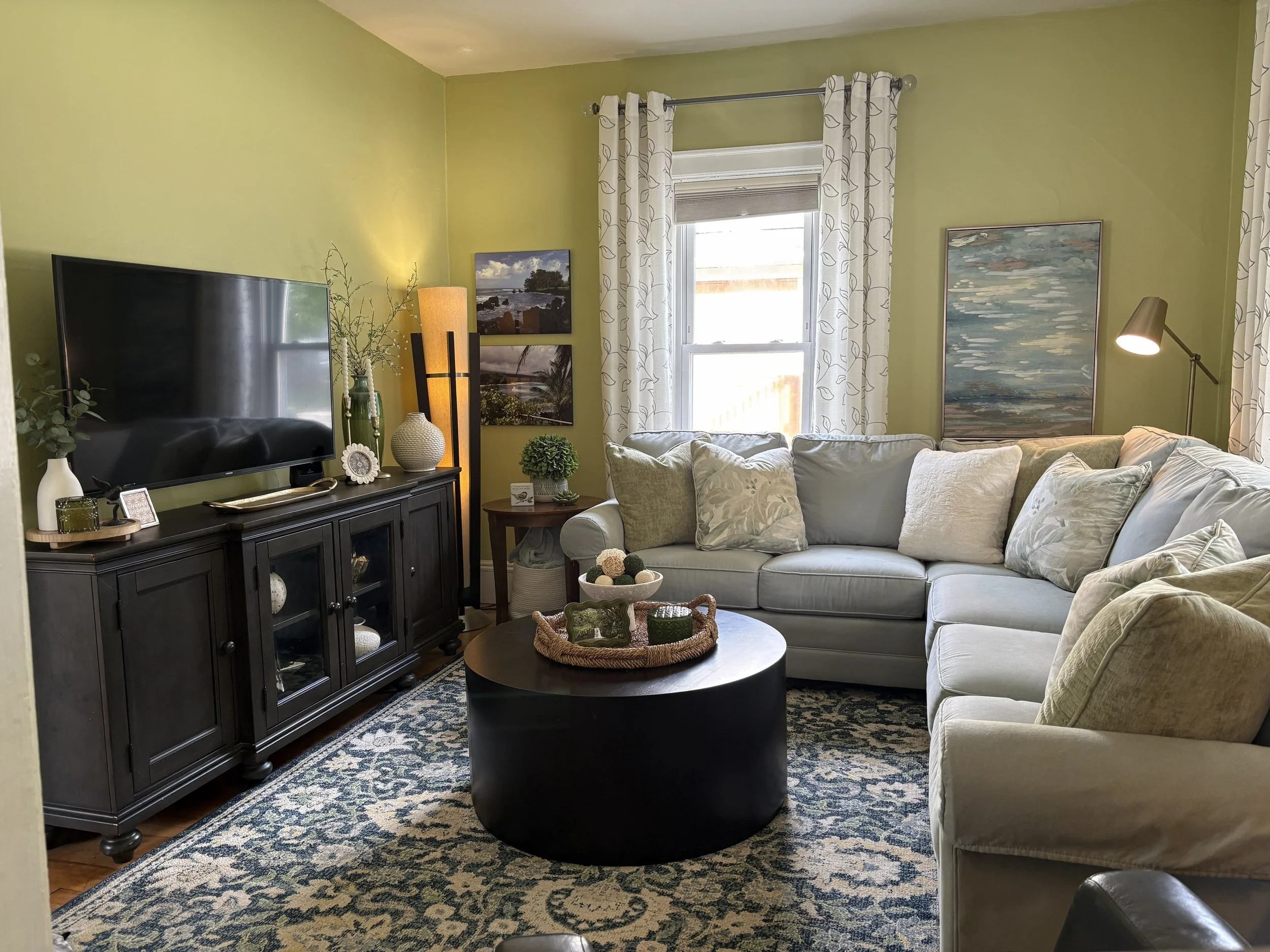 Living room with green walls, beige sofa, black coffee table, TV on black cabinet, decorative items, paintings, window with white curtains.