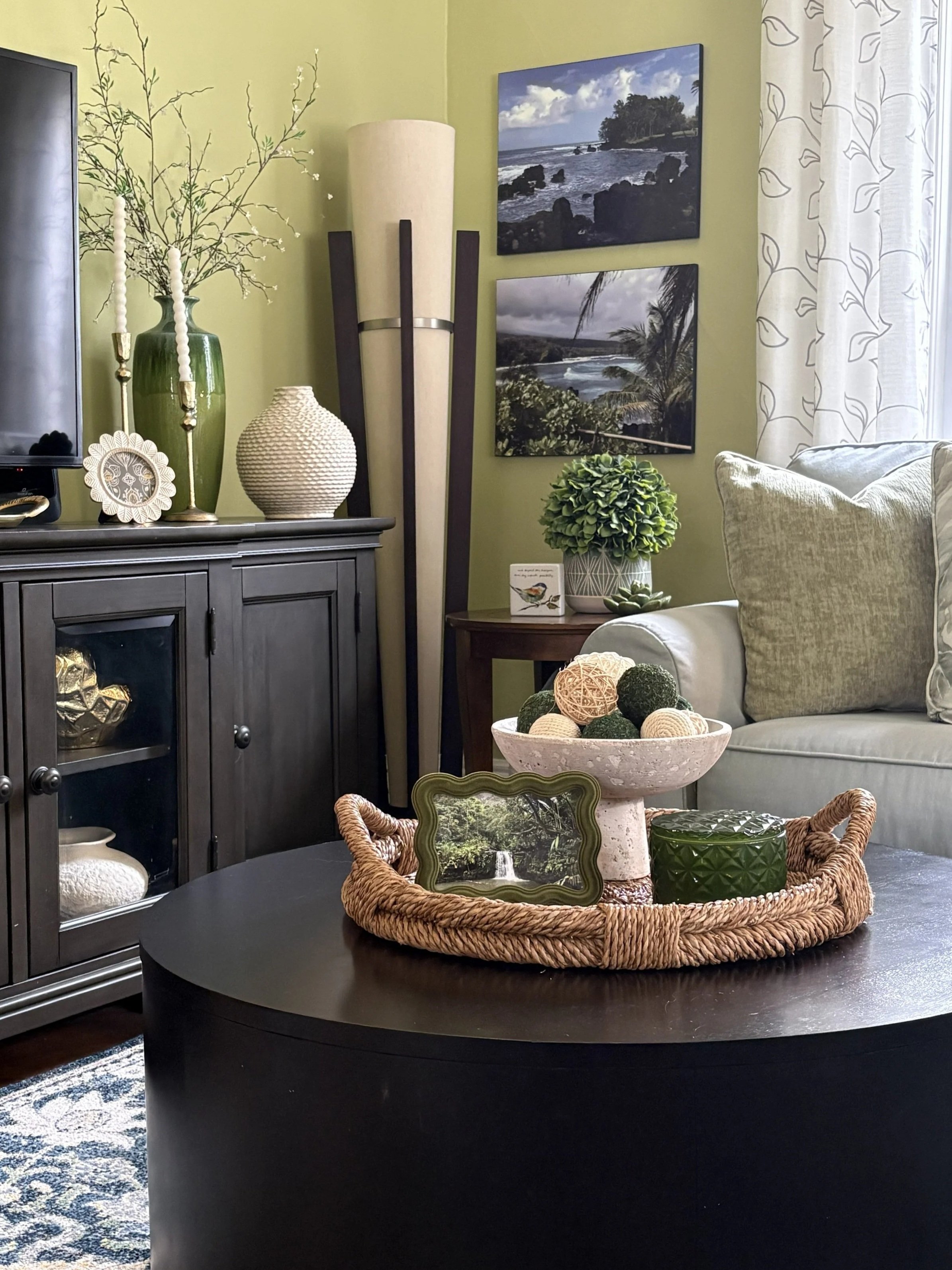 Living room with a black coffee table, a gray cabinet with decorative items, a beige couch with pillows, and framed landscape photographs on the wall.