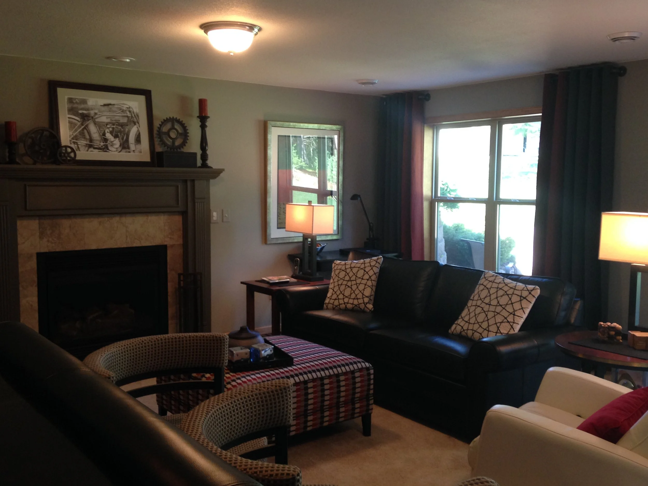 Living room with a fireplace, framed black and white bicycle artwork, window with blue curtains, various chairs, and a lamp on a side table.