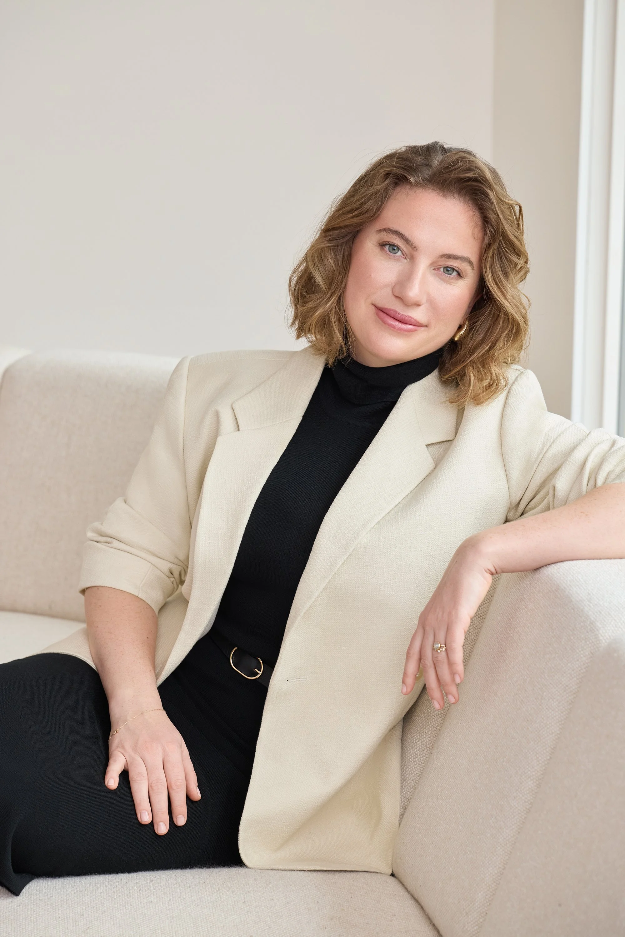 A woman with shoulder-length wavy blonde hair and blue eyes sitting on a beige couch, wearing a black turtleneck, cream-colored blazer, and black pants, with rings and a gold bracelet, smiling at the camera.