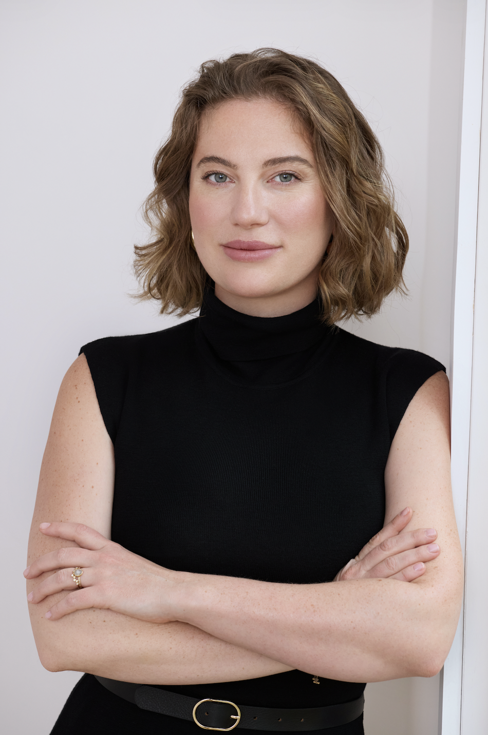 Portrait of a woman with light skin, short wavy brown hair, wearing a black sleeveless turtleneck top, standing against a light background with her arms crossed.