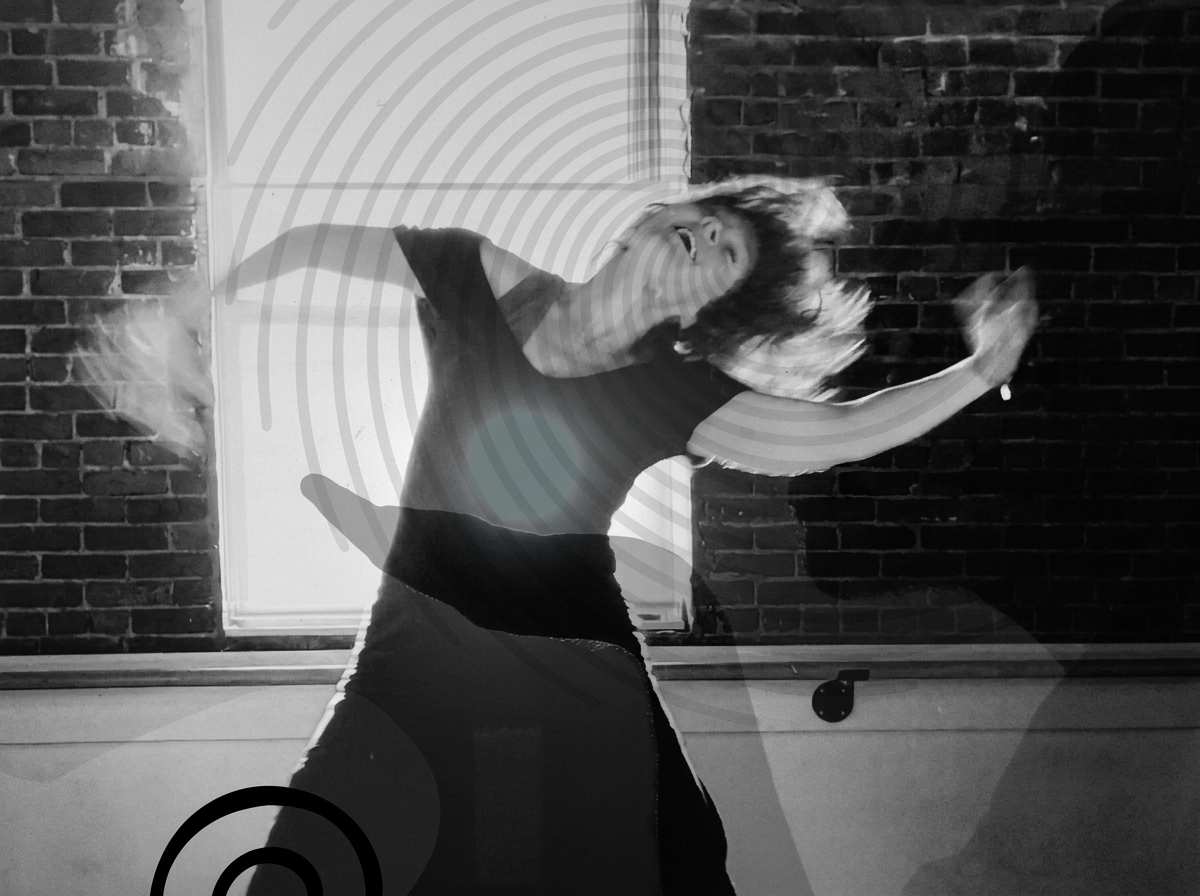 A woman dancing or moving energetically near a brick wall and window, with her hair flying and one arm raised.