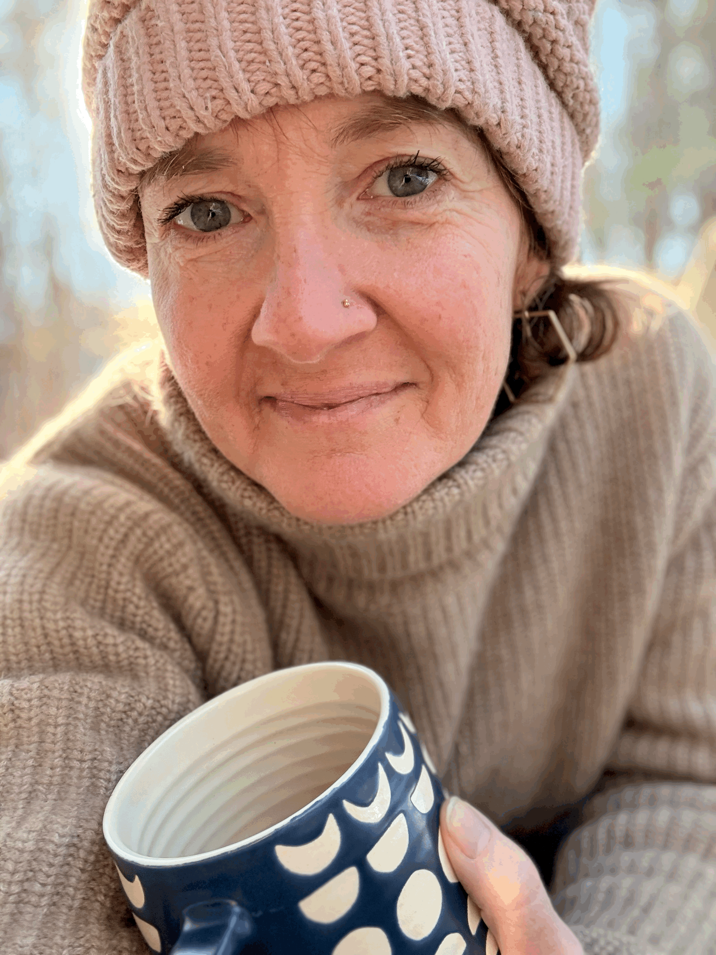 A woman wearing a pink knit beanie and a beige sweater, holding a blue ceramic mug with white polka dots, outdoors with blurred trees in the background.