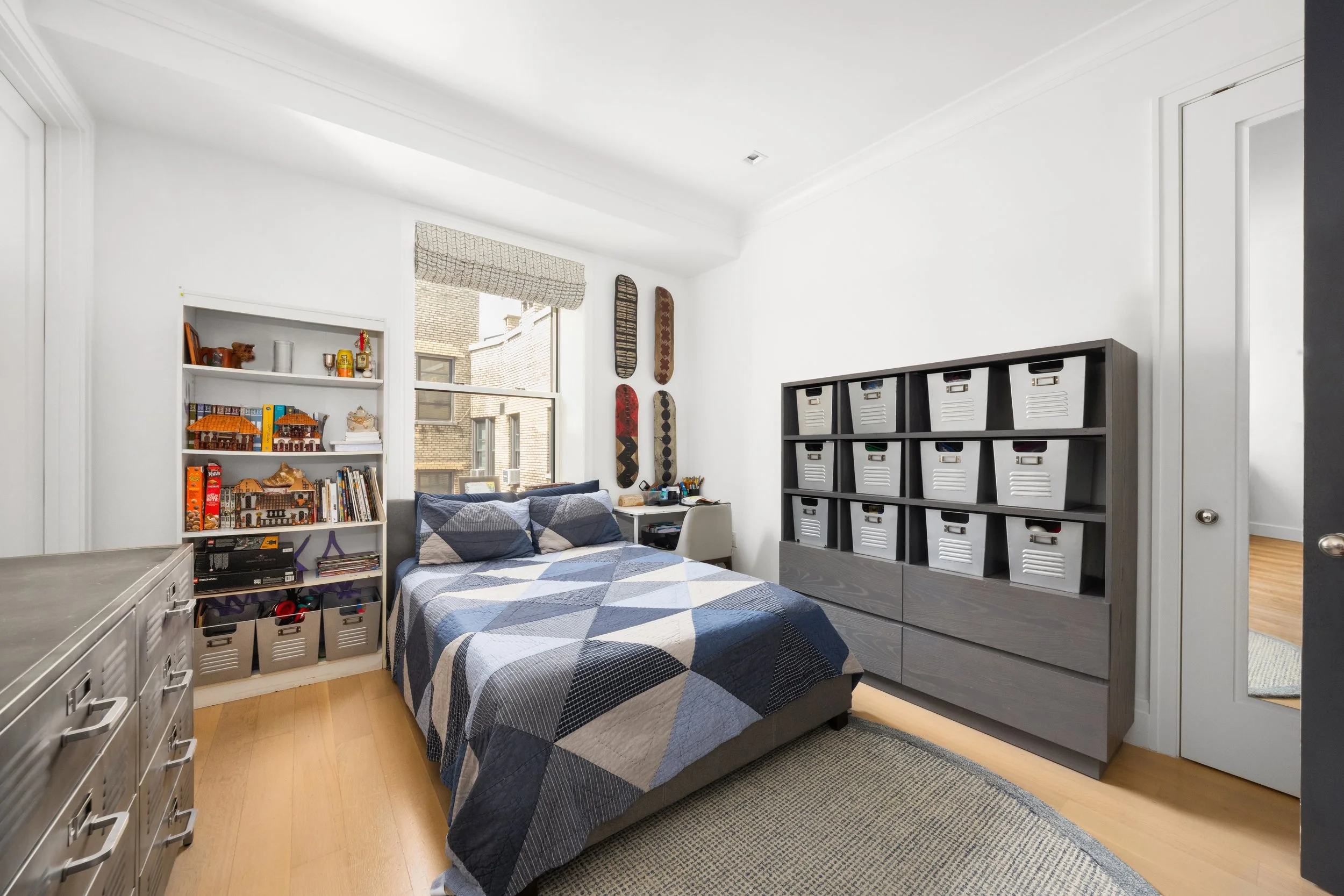 A cozy bedroom with a bed covered in a black, white, and gray geometric quilt, flanked by white shelves filled with books and decorations, with large window letting in natural light, and a tall gray storage unit with labeled baskets.