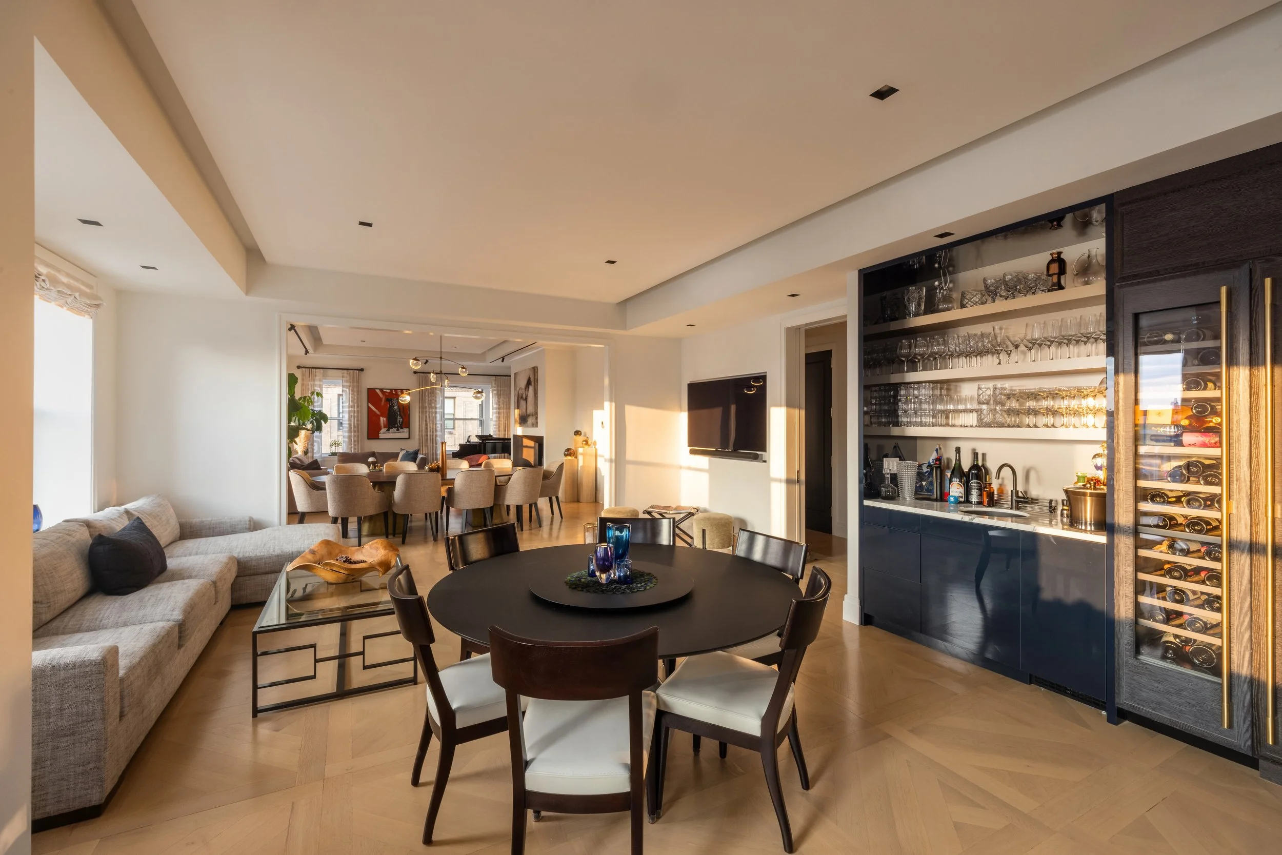 Interior of a modern, open-concept living and dining area with a bar, glassware, and wine fridge, illuminated by sunlight, featuring a beige sofa, round dining table, and wooden floors.