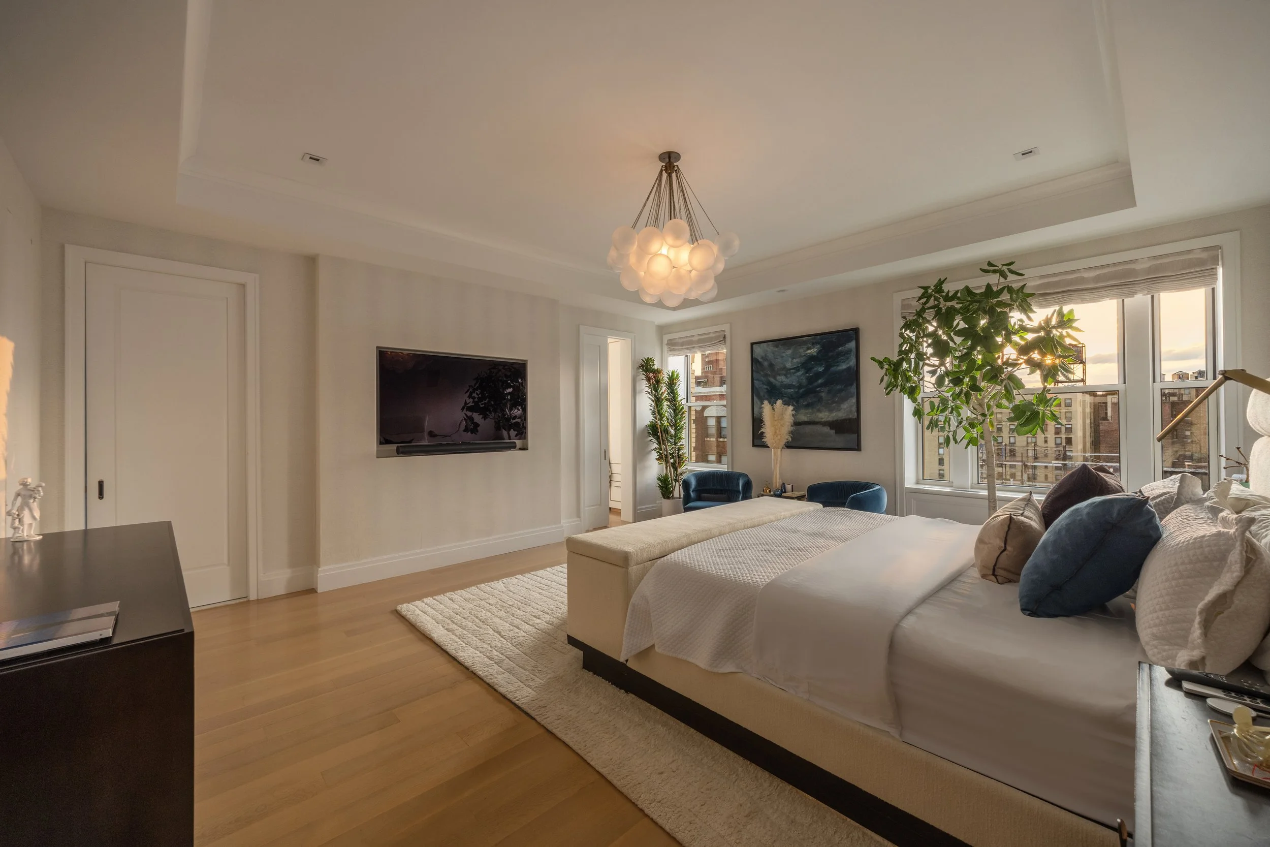 Living room with a white sofa, navy blue chairs, a large window, a TV, a chandelier, and decorative plants in a city apartment.