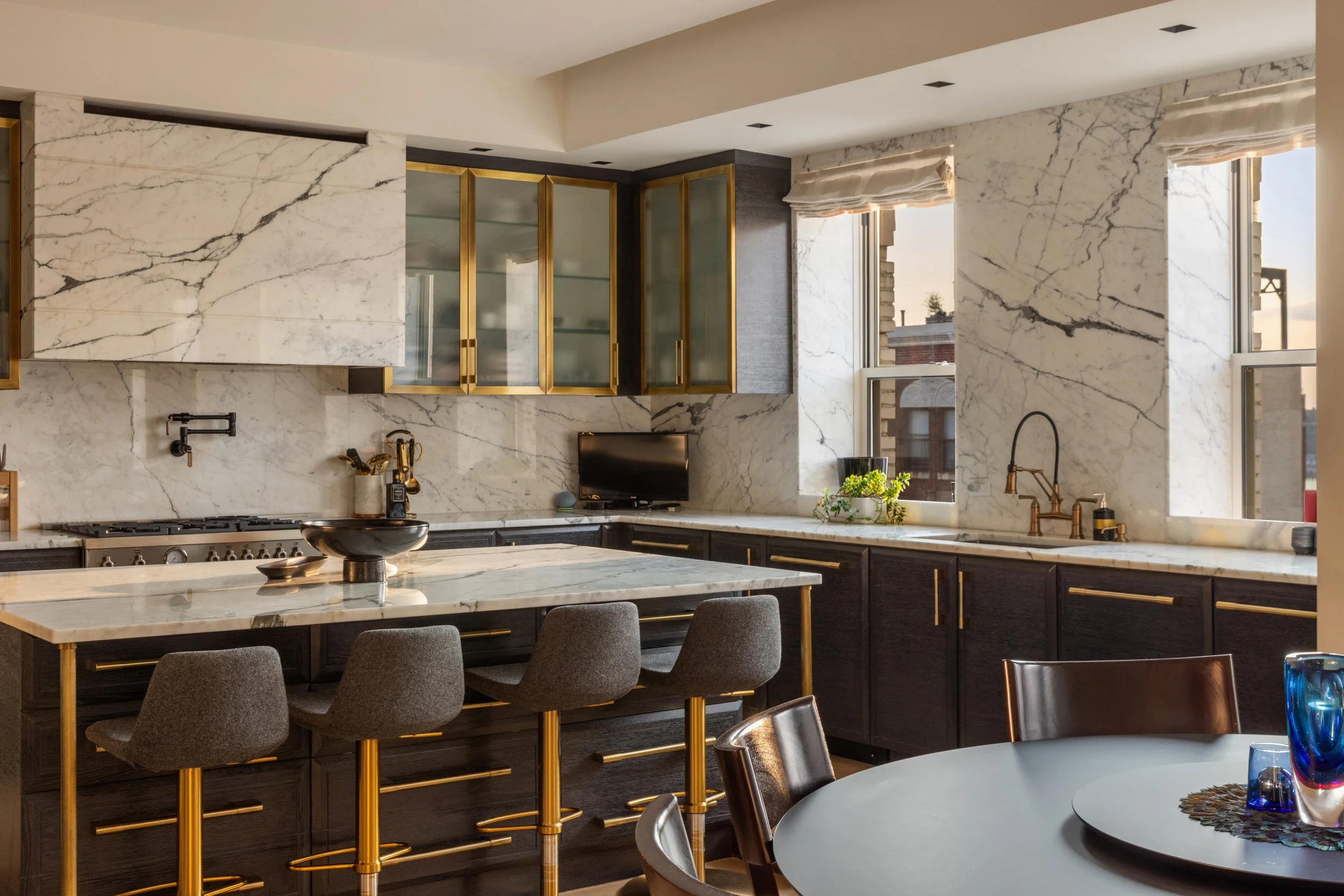 Modern kitchen with white marble countertops, dark cabinets with gold handles, glass-front cabinets with gold trim, and large windows letting in natural light.
