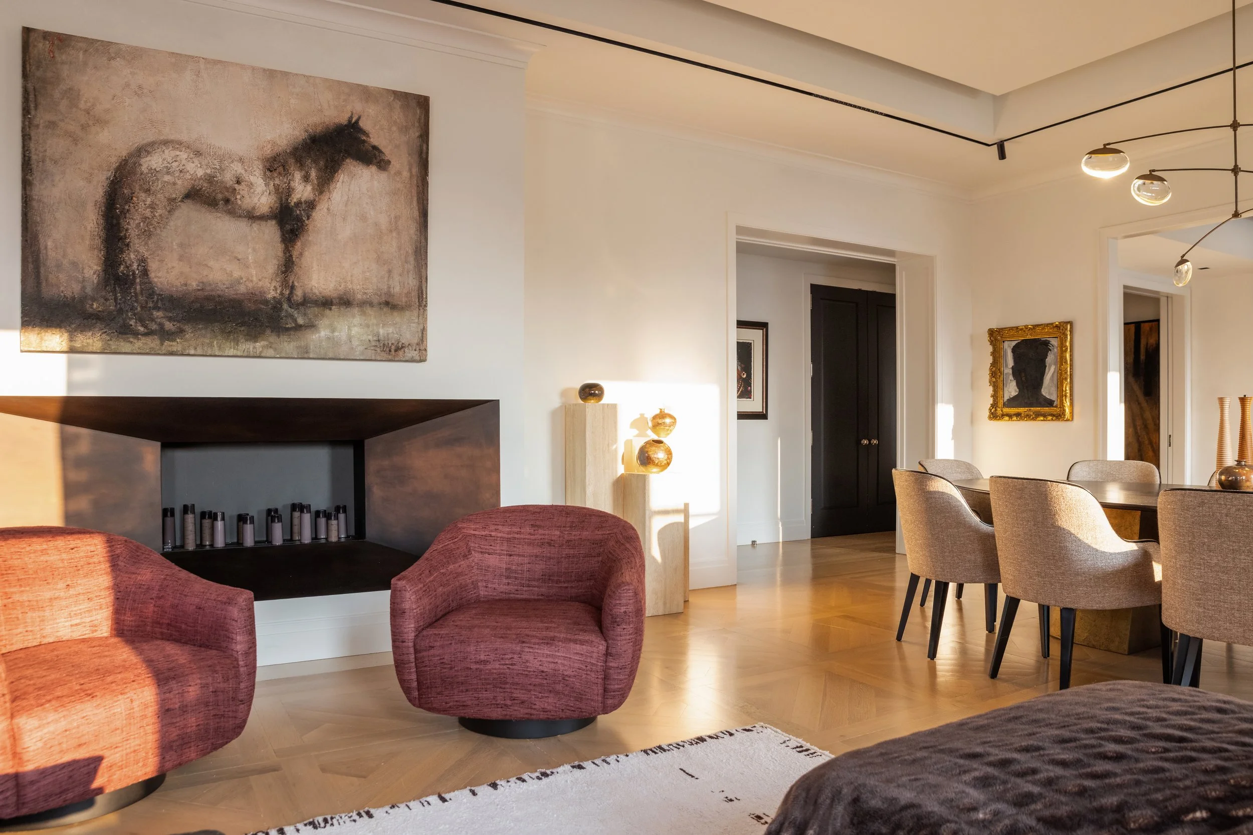 Living room with pink armchairs, a painting of a horse on the wall, and a dining area with chairs and a pendant light. Sunlight streams in through the windows, illuminating the room.
