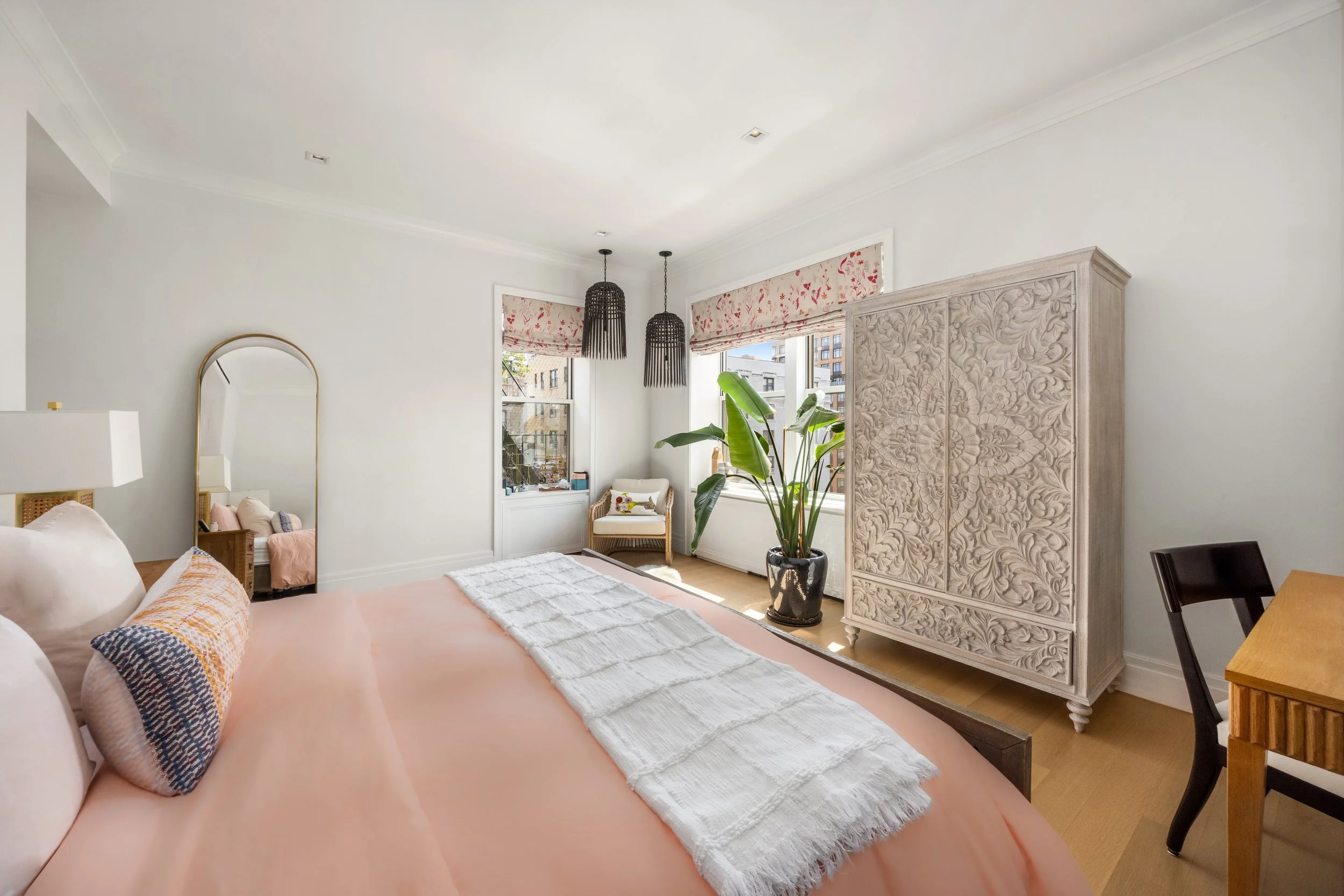 A bright, airy bedroom with a large bed, decorative pillows, a mirror, a potted plant, and a tall carved wooden wardrobe, with large windows and hanging pendant lights.