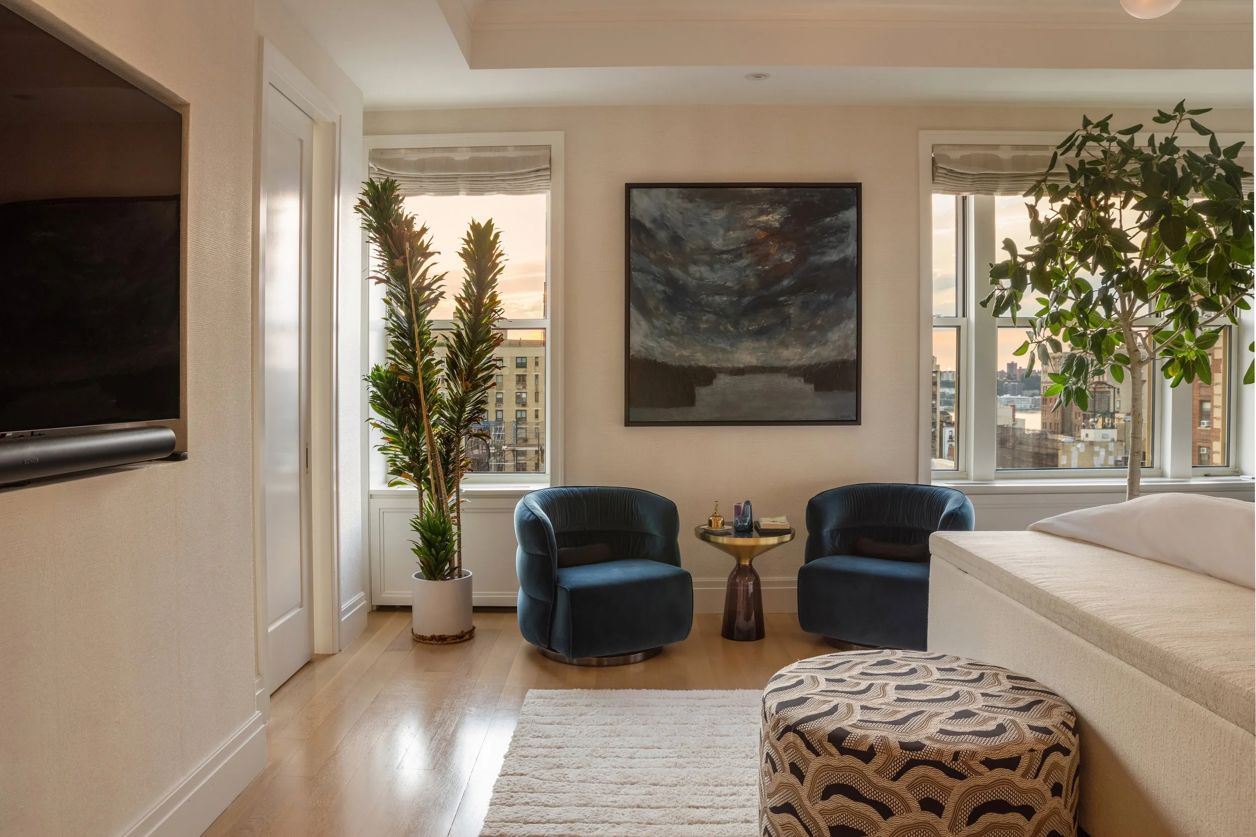 A modern living room with large windows showing city skyline views, two black armchairs around a small gold and black side table, a potted plant, a framed abstract painting, and a bed partially visible on the right.