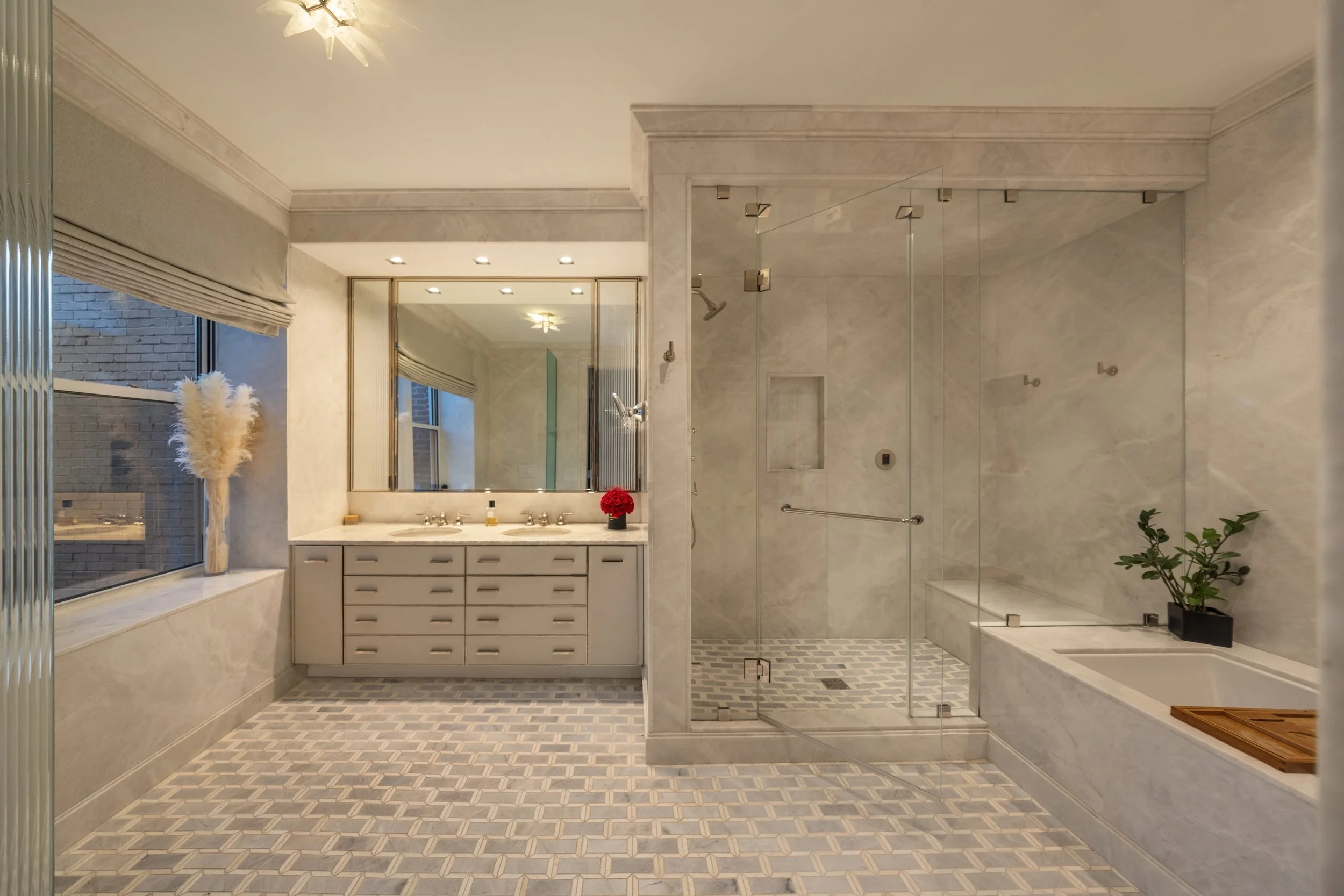 A luxurious bathroom with a large vanity featuring two sinks, a big mirror, and a window with a blinds. There is a floral plant on the window sill and a red flower in a vase on the vanity. To the right, there is a glass-enclosed shower with a marble 
