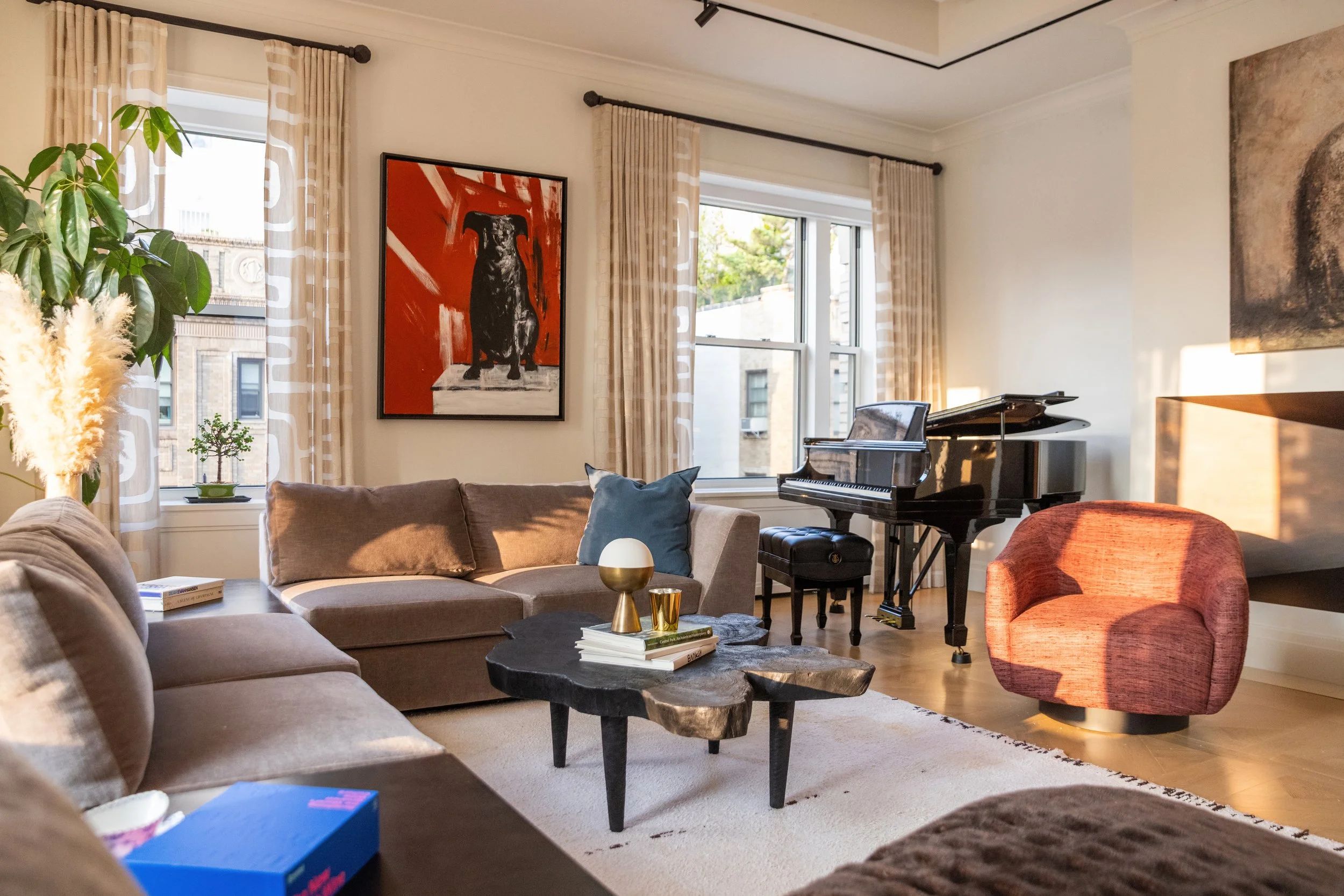 A living room with large windows, beige curtains, potted plants, artwork on the walls, a beige sofa with pillows, a black coffee table with books and decorative items, a grand piano, and a red upholstered armchair.