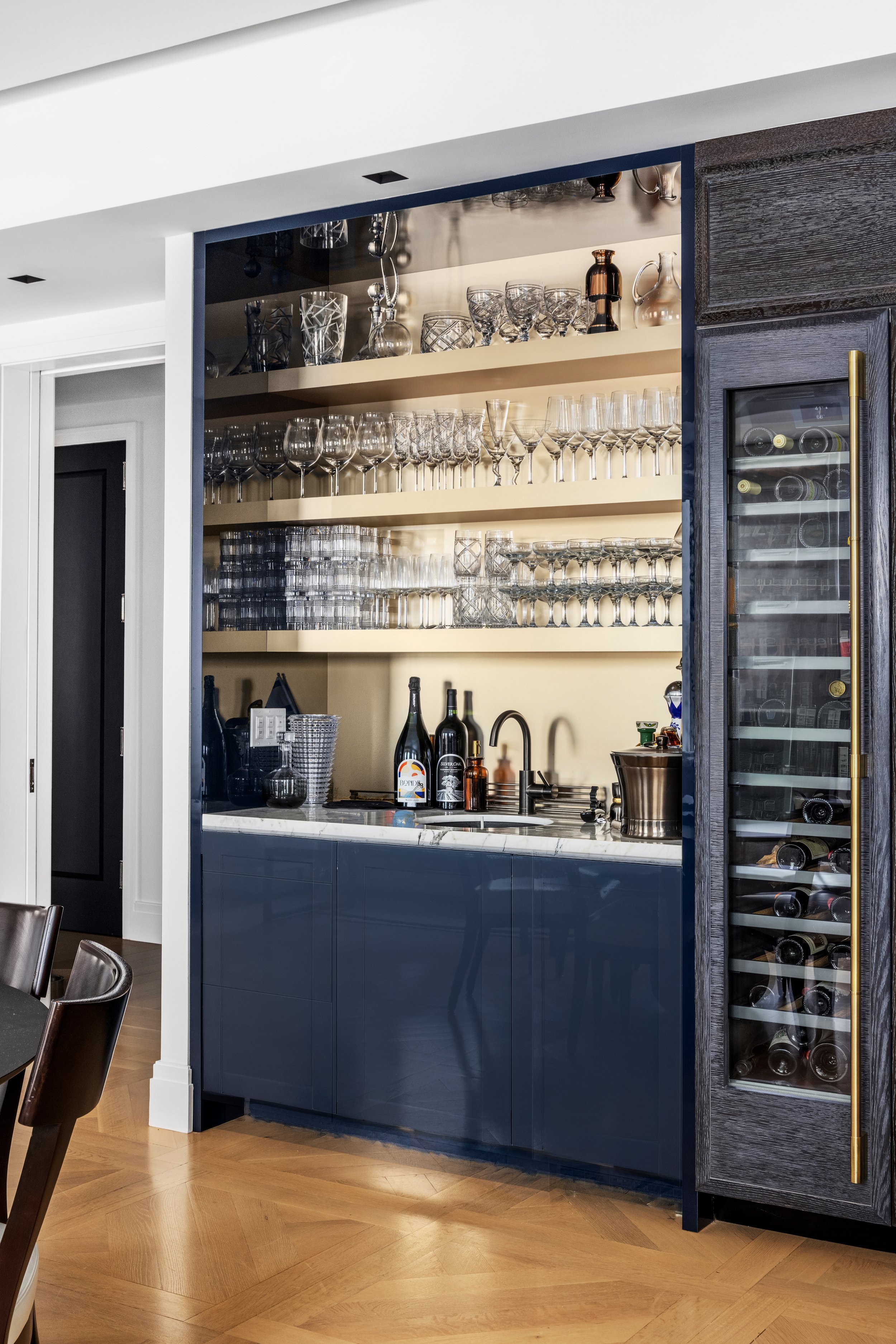 Open kitchen cabinet with shelves of glassware, a wine fridge, and bar tools on countertop in a modern kitchen.