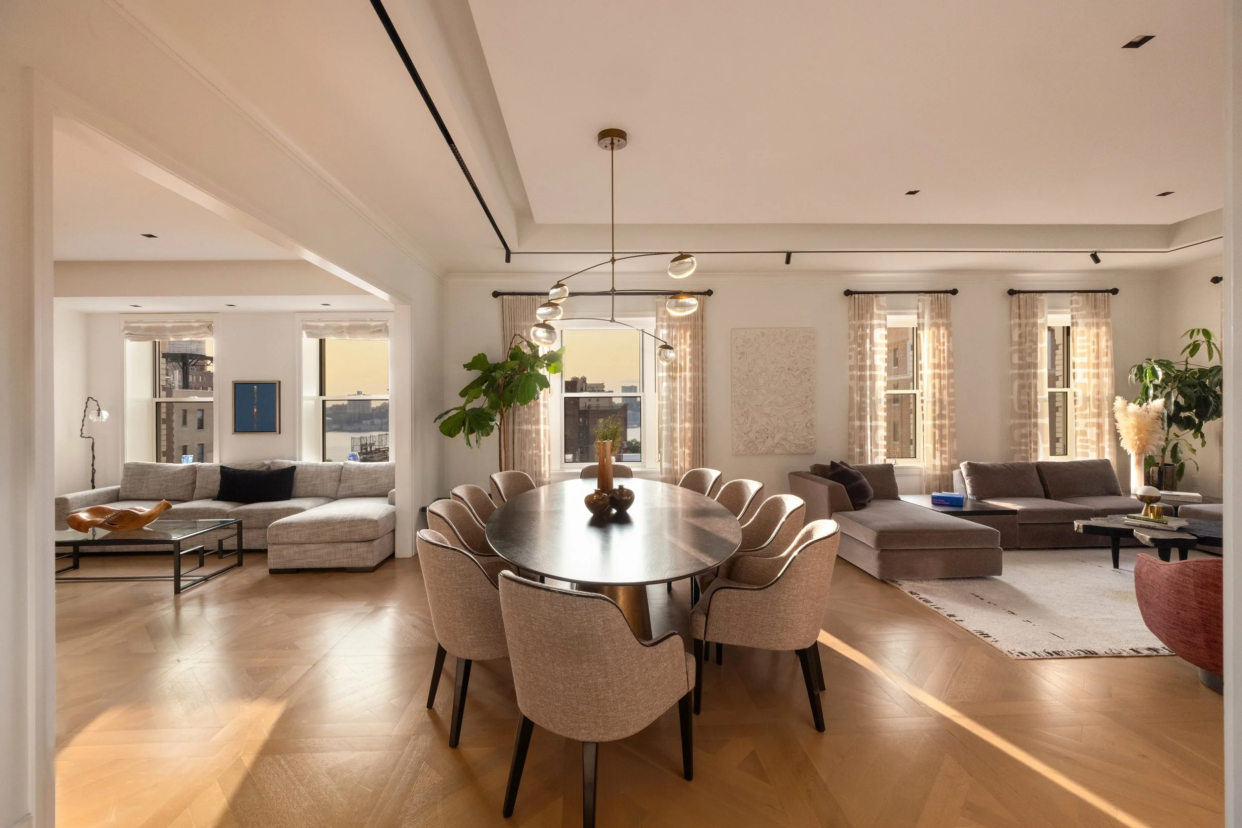 Spacious modern living room and dining area with large windows, beige curtains, several sofas, a round dining table with beige chairs, and decorative items including plants and artwork.