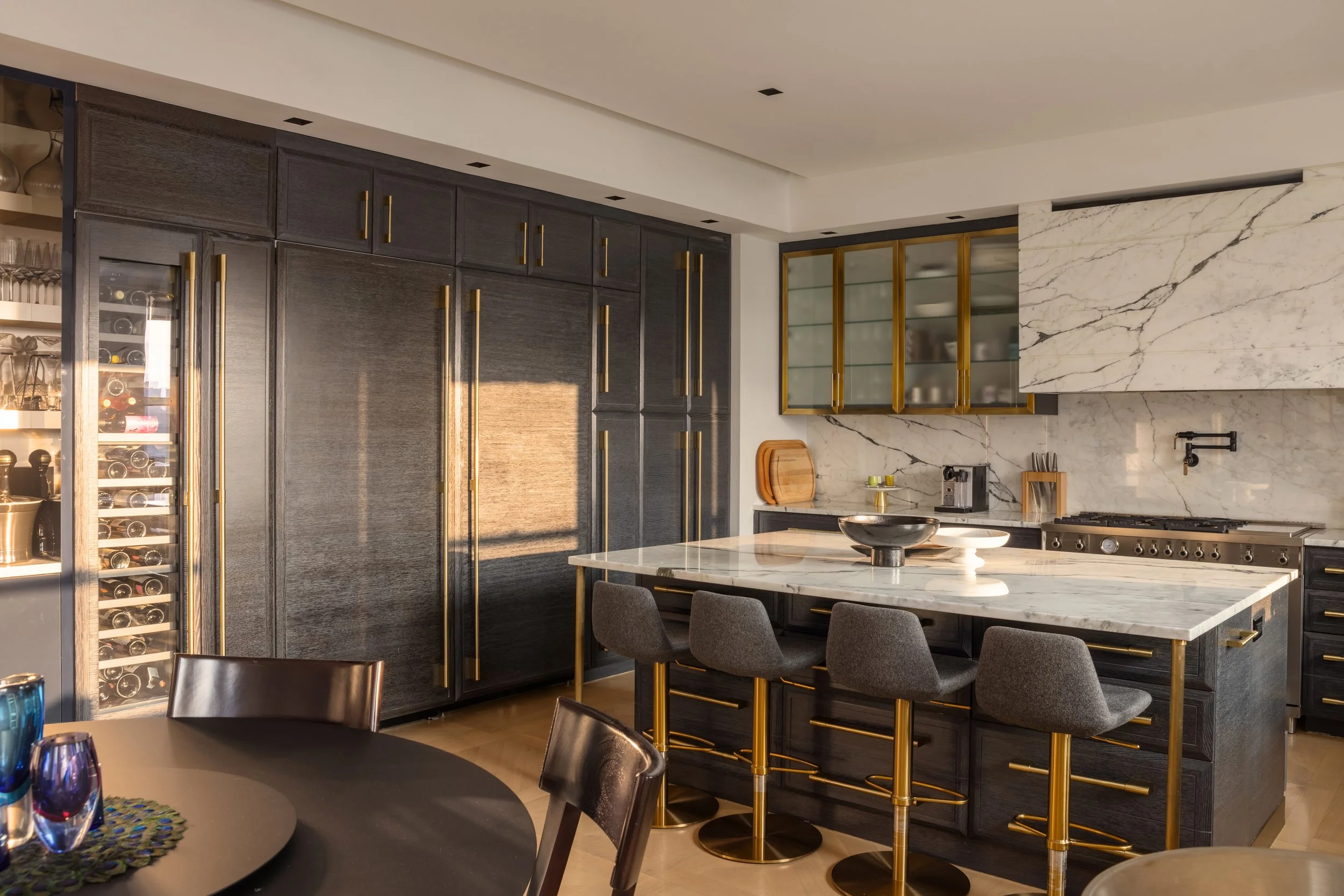 Modern kitchen with black cabinetry, gold handles, marble countertops, and a built-in wine fridge. Central island with a marble top and gray chairs, and shelves with glassware and dishes.