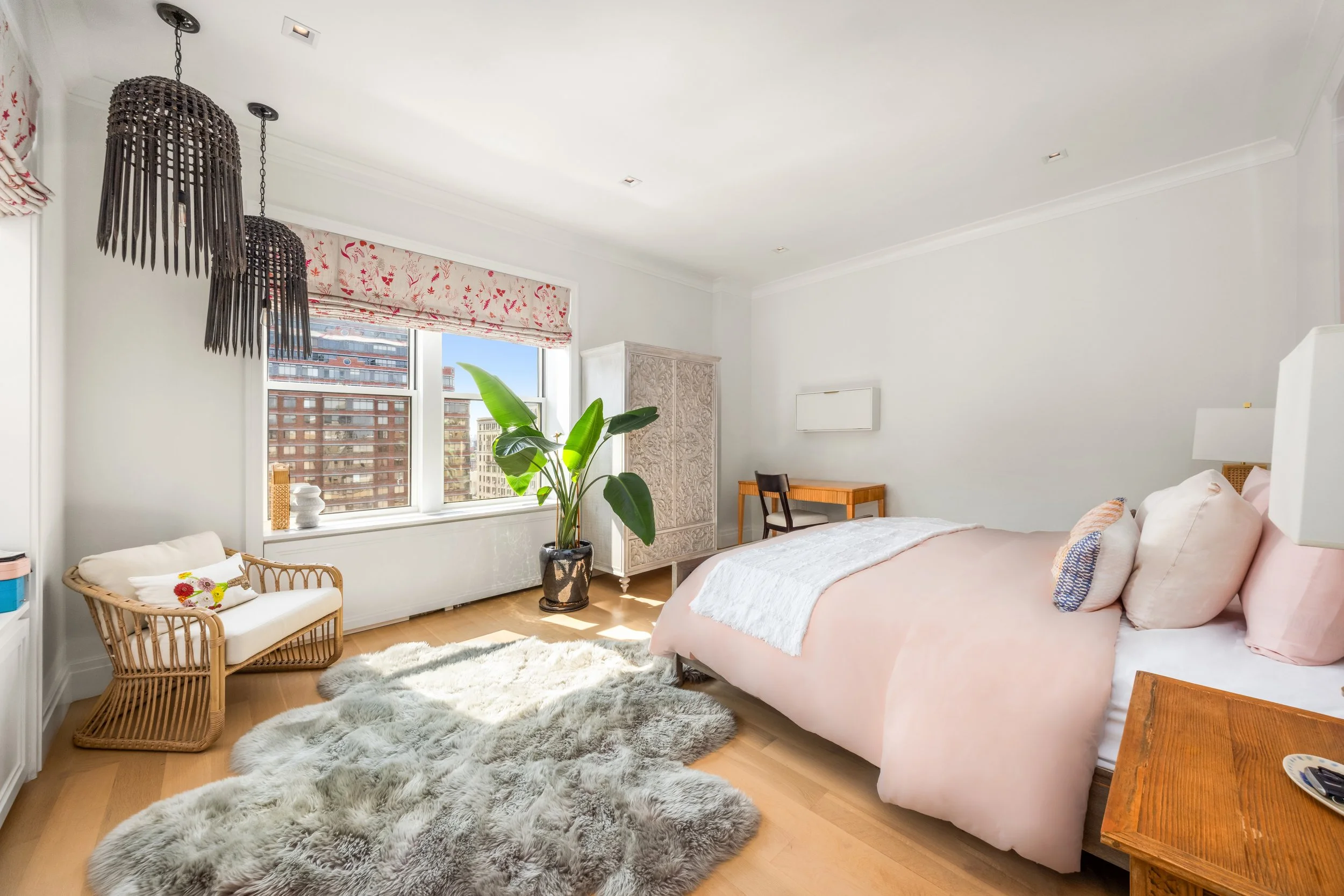 A bedroom with a large bed, pink bedding, and pillows. A window with white blinds and floral curtains, a plant on a stand, a wicker bench with white pillows, a wooden table, and a white cabinet. Plush gray rug on light wooden floor.
