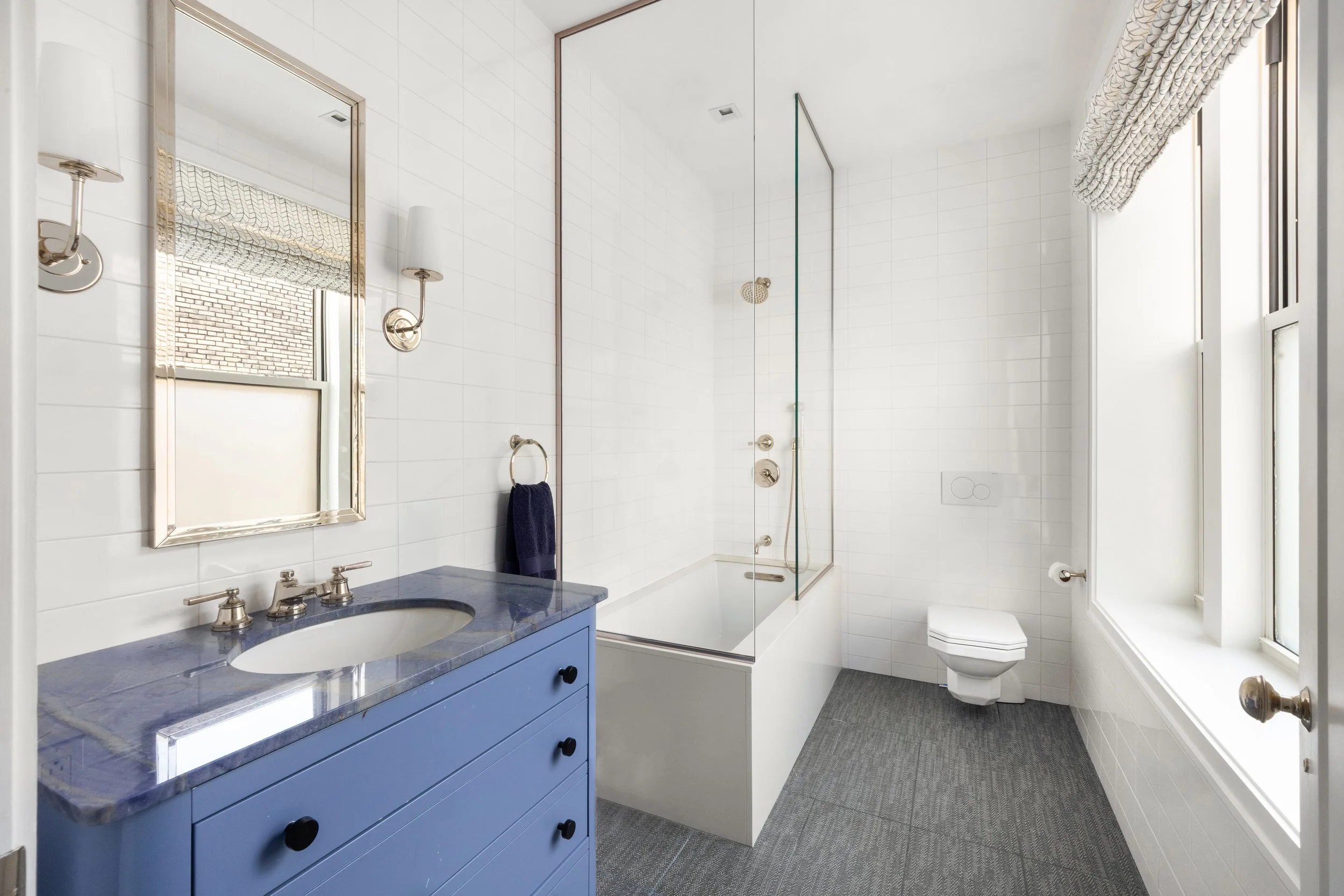 A clean, modern bathroom with white tiled walls, a blue vanity with a marble countertop, a mirror, a sink, a wall-mounted lamp, a glass-enclosed shower, and a white toilet near a window with a patterned curtain.