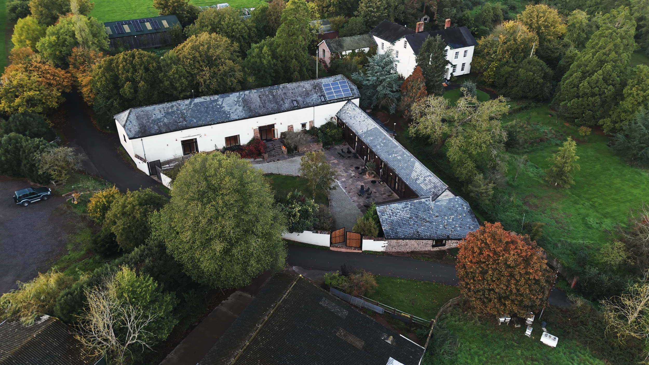 Aerial view of a historic estate with slate-roofed buildings, solar panels, landscaped gardens, and surrounding woodland.