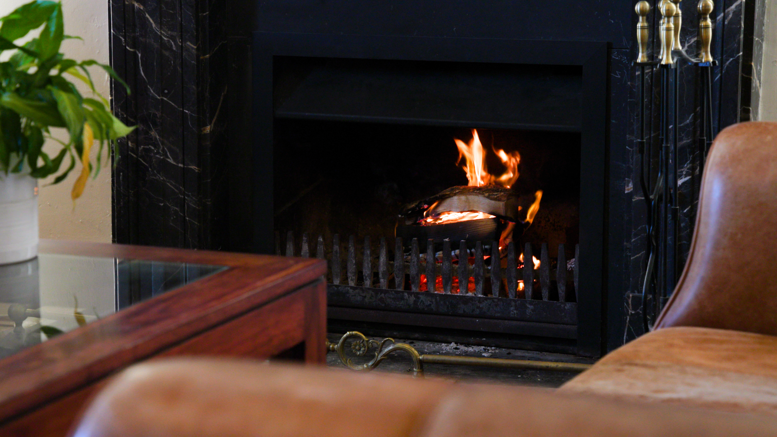 Cozy living room with a crackling fire in a black marble fireplace, fireplace tools, a houseplant, and leather chairs