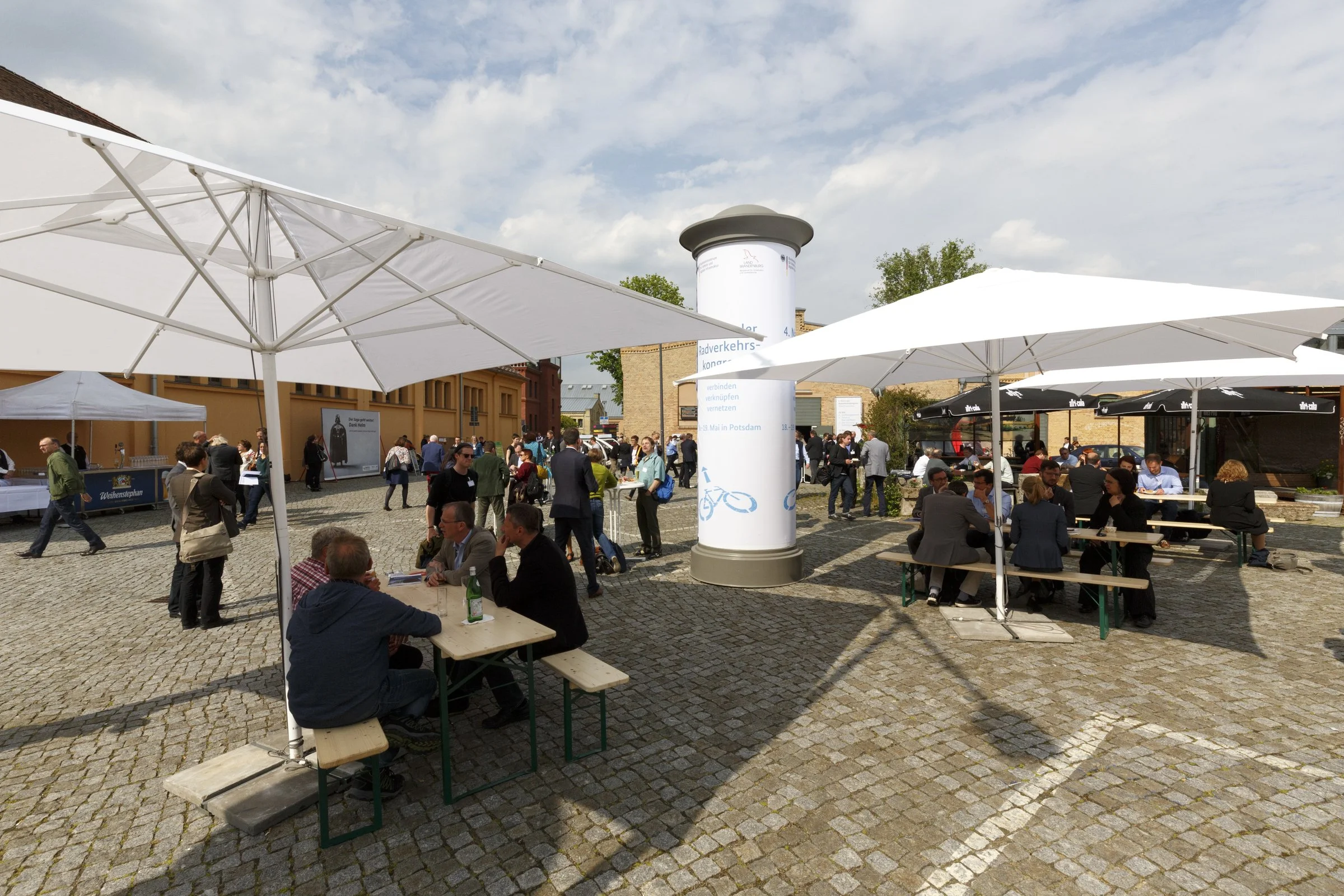 Menschen versammelt auf einer offenen cobblestone Plaza, unter weißen Sonnenschirmen, bei einer Veranstaltung, mit mehreren Sitzgelegenheiten, die an einem warmen Tag stattfinden.