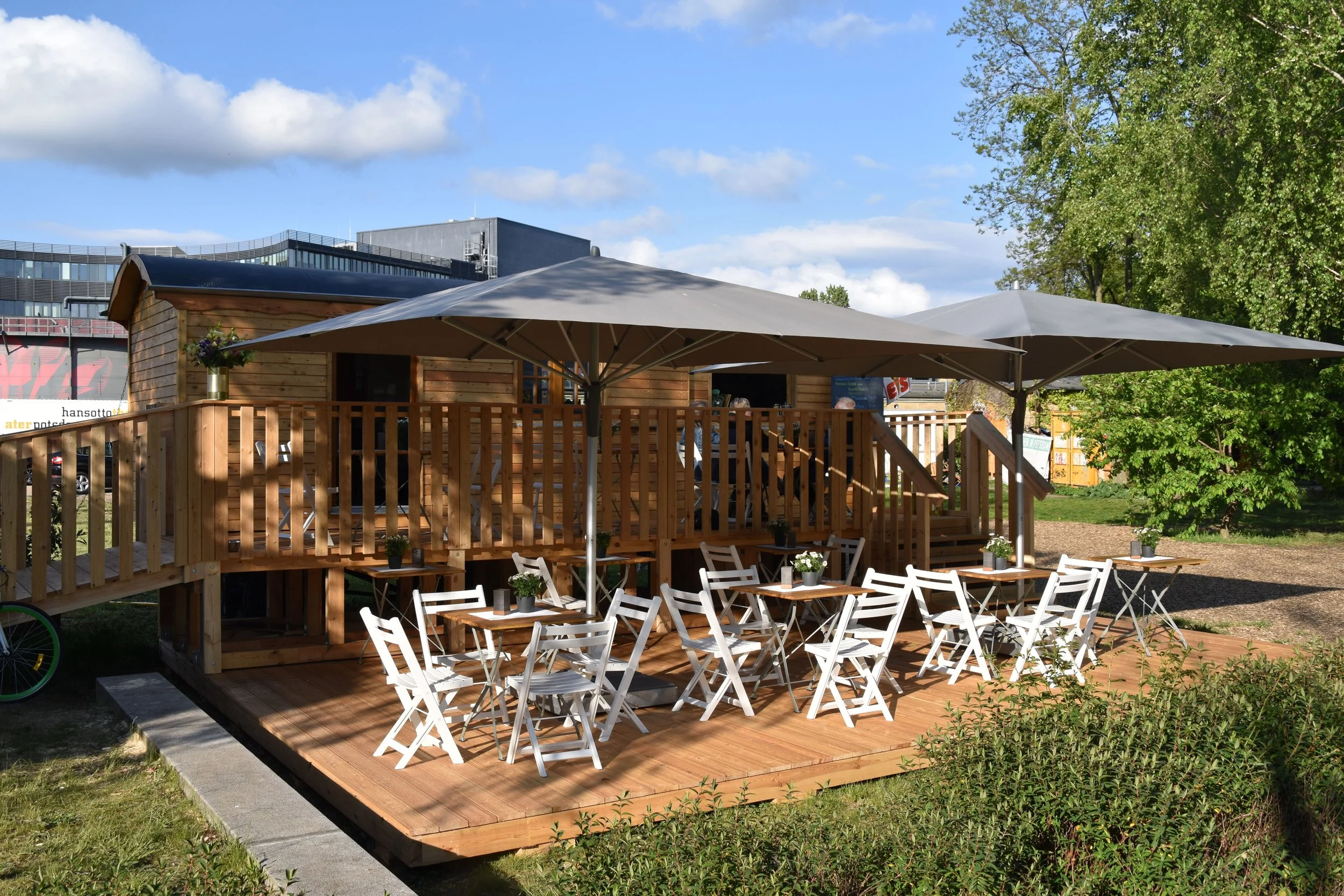 Ein im Freien stehender Holzzaun mit einem Bistro im Vordergrund, dekoriert mit kleinen Pflanzen auf den Tischen. Im Hintergrund befinden sich große Sonnenschirme, ein Gebäude mit großen Fenstern und Bäumen unter einem blauen Himmel mit einigen Wolken.