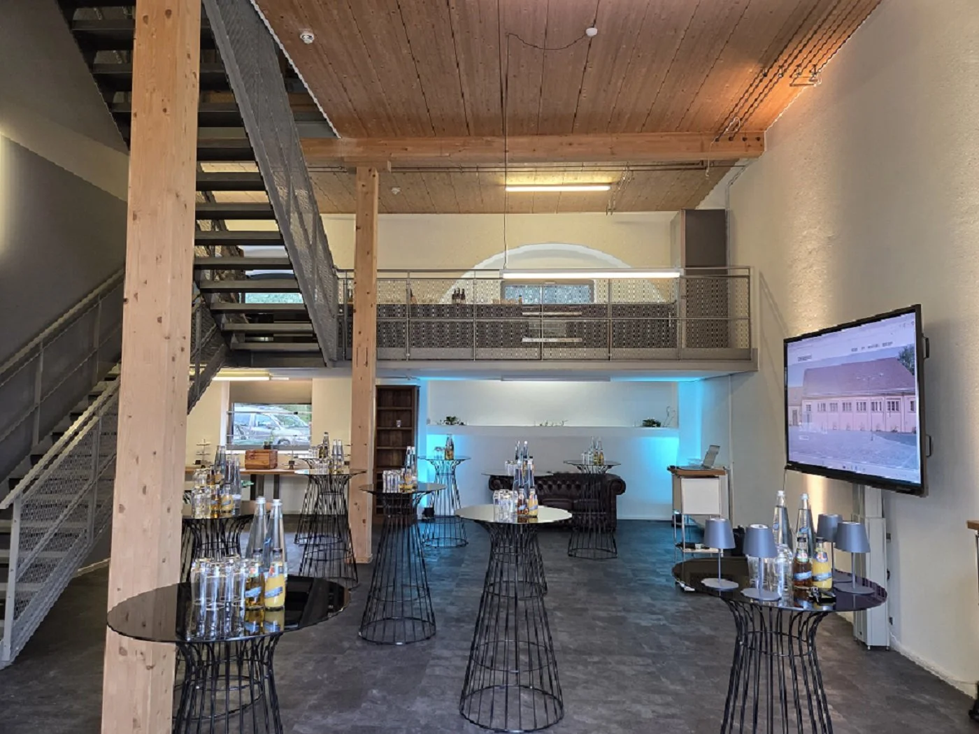 Innenansicht eines modernen Eventraums mit schwarzen Stehtischen, Wasserflaschen und Gläsern, einer großen Wandbildschirm, einem Sofa und einer neuen Lounge. Holzdecke, Metalltreppe und eine zweite Etage sind sichtbar.