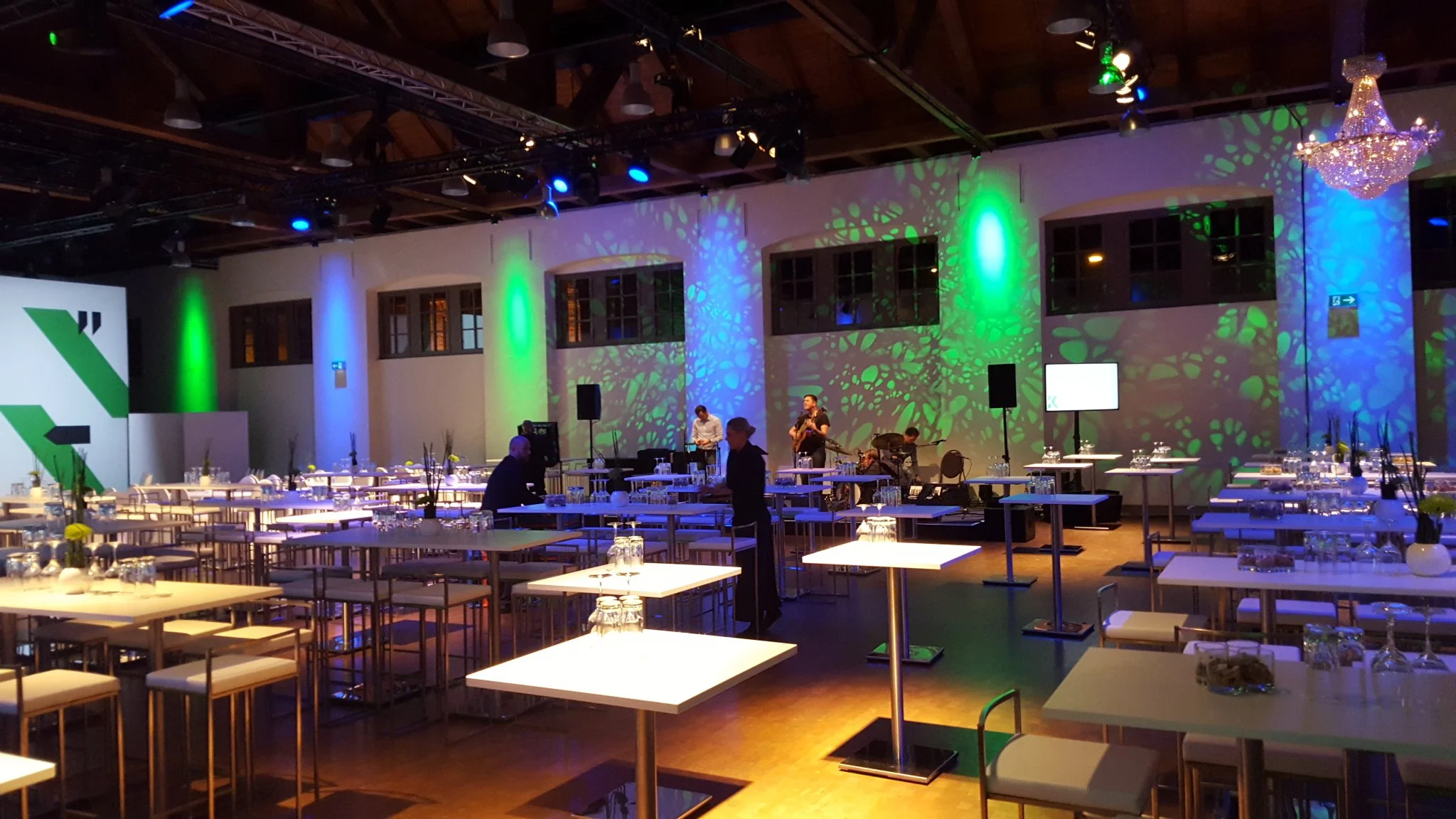 Innenaufnahme eines festlich gedeckten Raumes mit Tischen, einer Bühne mit Live-Musikern und bunten Lichteffekten, eine elegante Kronleuchter und mehrere Fenster.