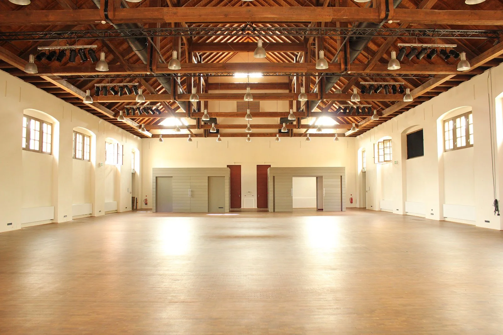 Ein großer, leerer Saal mit Holzboden, hohen weißen Wänden, mehreren hohen Fenstern, einer Holzbalkendecke und einer Deckenbeleuchtung, die bereits vorbereitet ist.