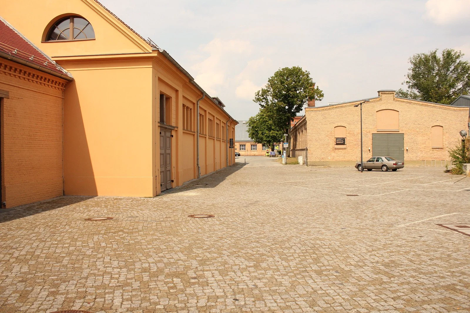 Ein leeres, gepflastertes Parkhaus mit einem grauen Auto auf der rechten Seite und mehreren Gebäuden mit alten Fassaden im Hintergrund, einige Bäume sind sichtbar.