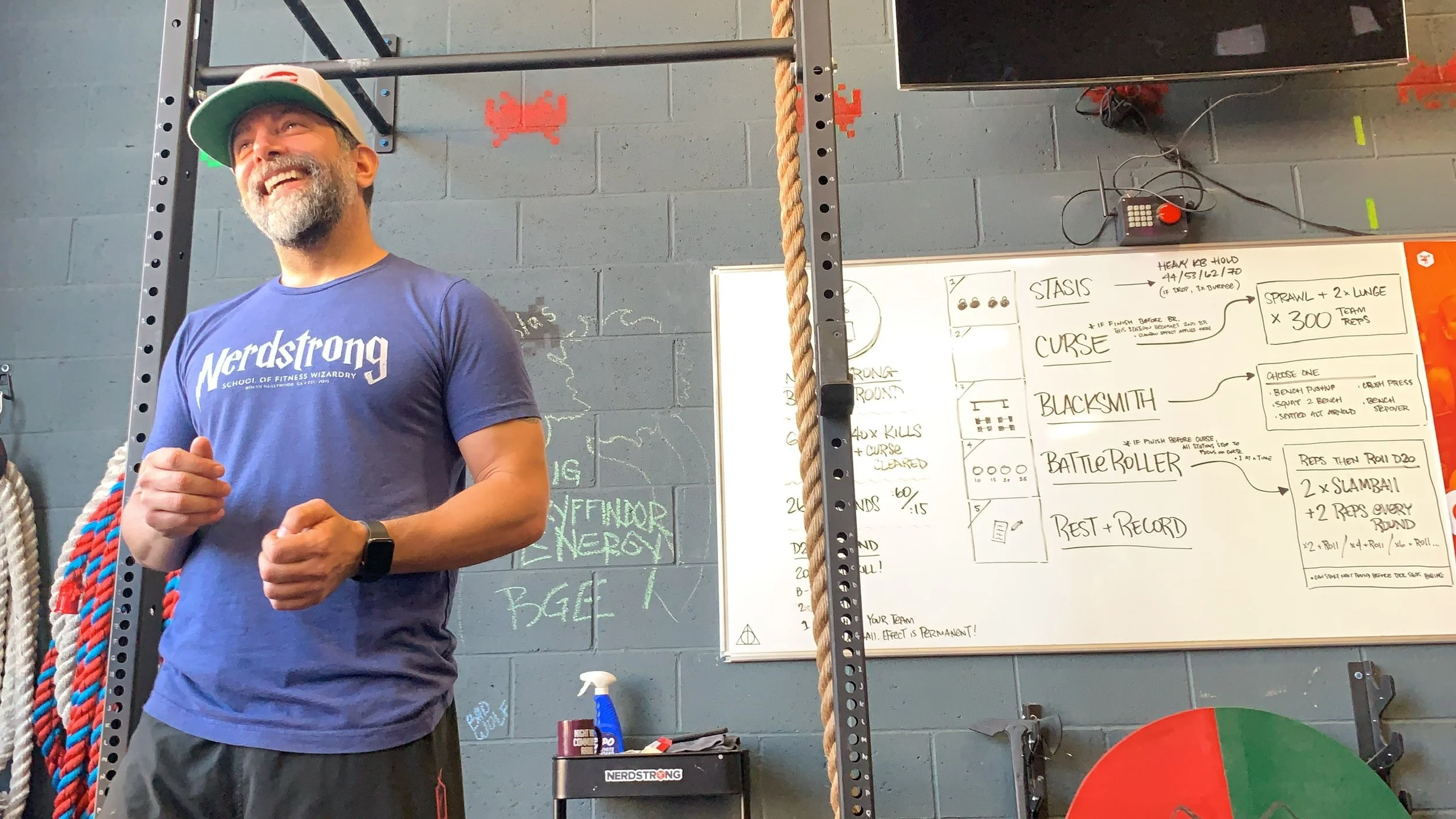 A man in a blue t-shirt standing in front of a whiteboard in a gym setting.