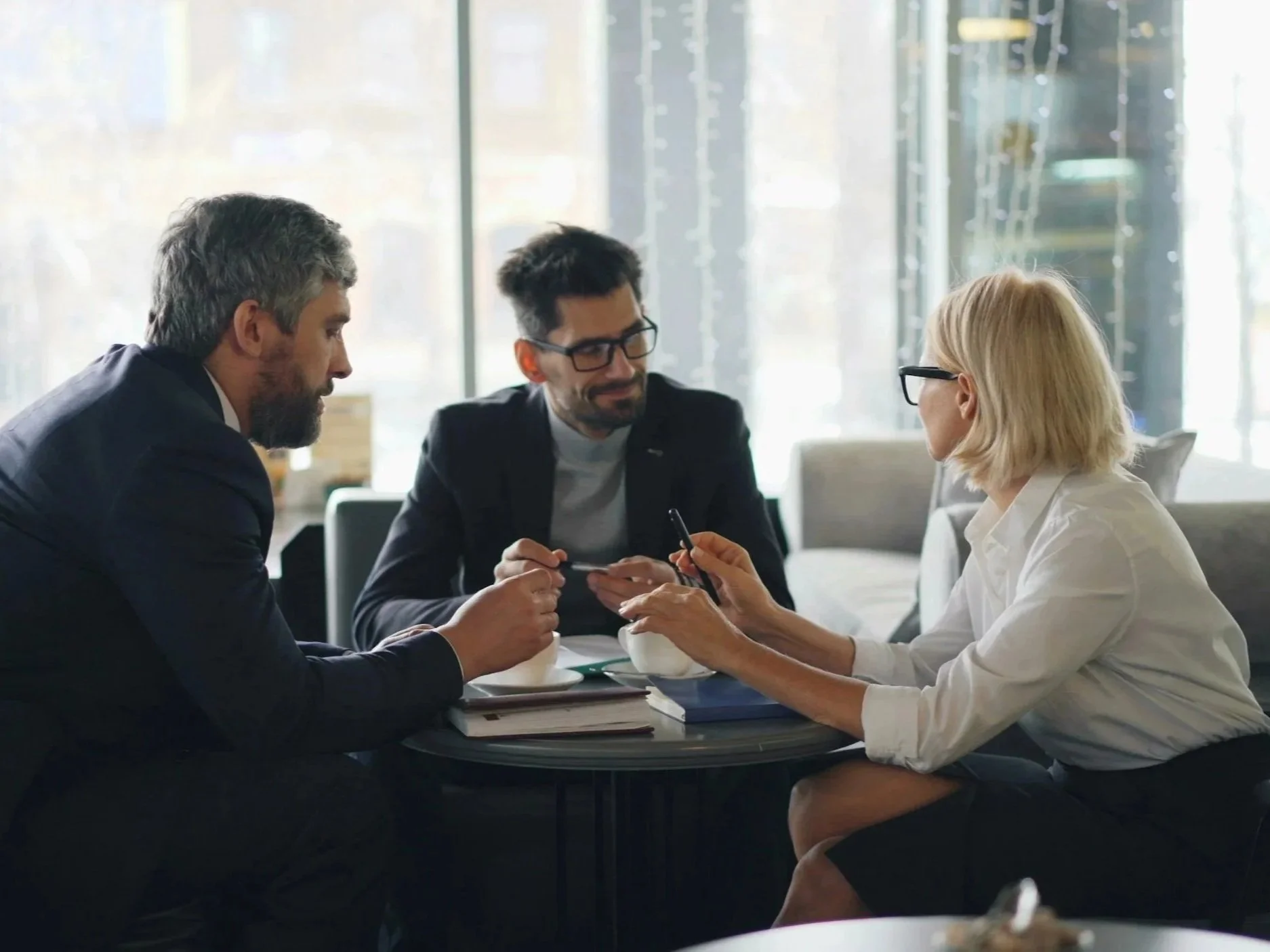 Three business professionals sitting at a table in a bright office, engaging in a discussion while looking at a smartphone, with two of them holding the device.