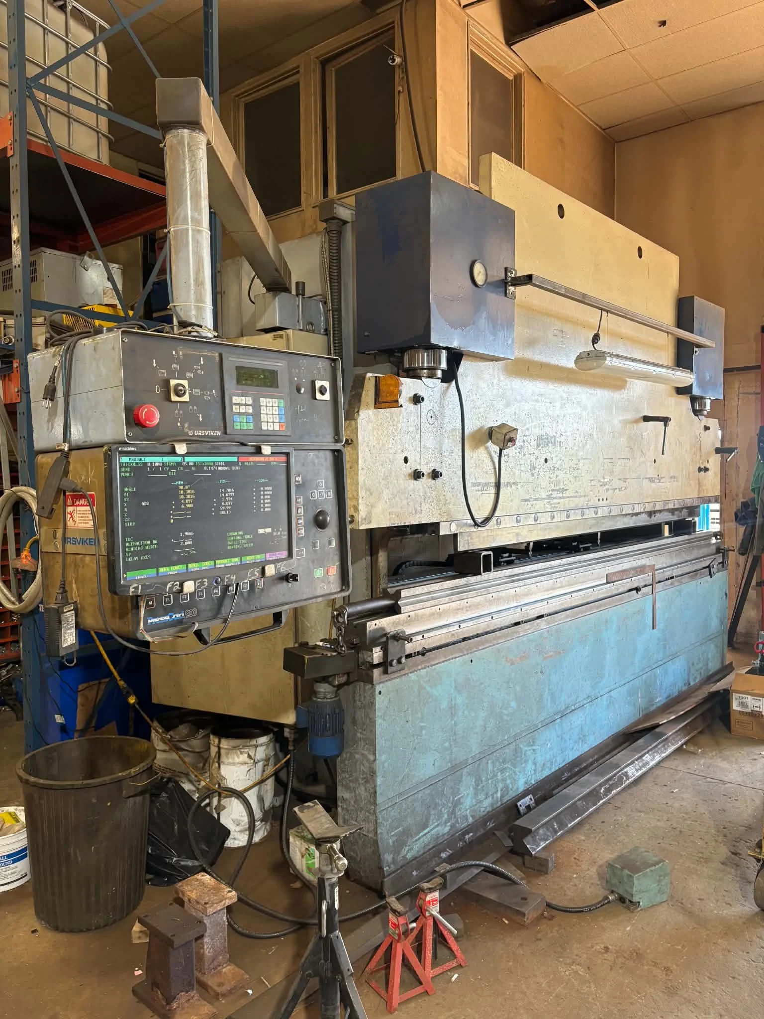 A workshop with a large machine or mill, possibly a CNC or lathe, on a workbench with a computer control panel. The workspace contains tools, brackets, and equipment, with some items on the floor and shelving.
