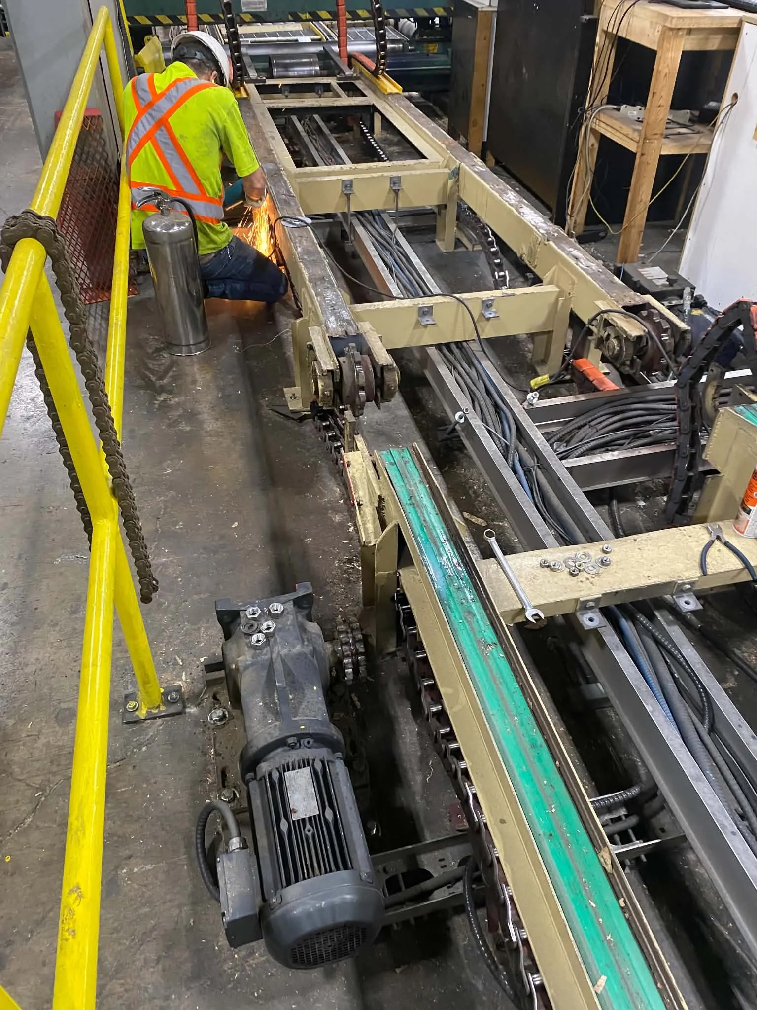 A worker with a safety vest and helmet is using a welding tool on a large industrial machine, which appears to be part of a manufacturing or assembly process, in a workshop or factory setting.