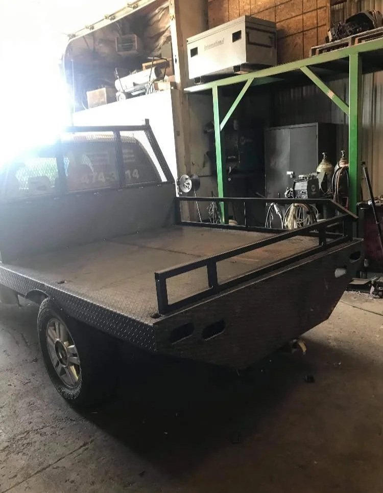 A black flatbed utility vehicle in a garage, with a metal railing on the front and a shelving unit mounted at the back, surrounded by various tools and equipment.