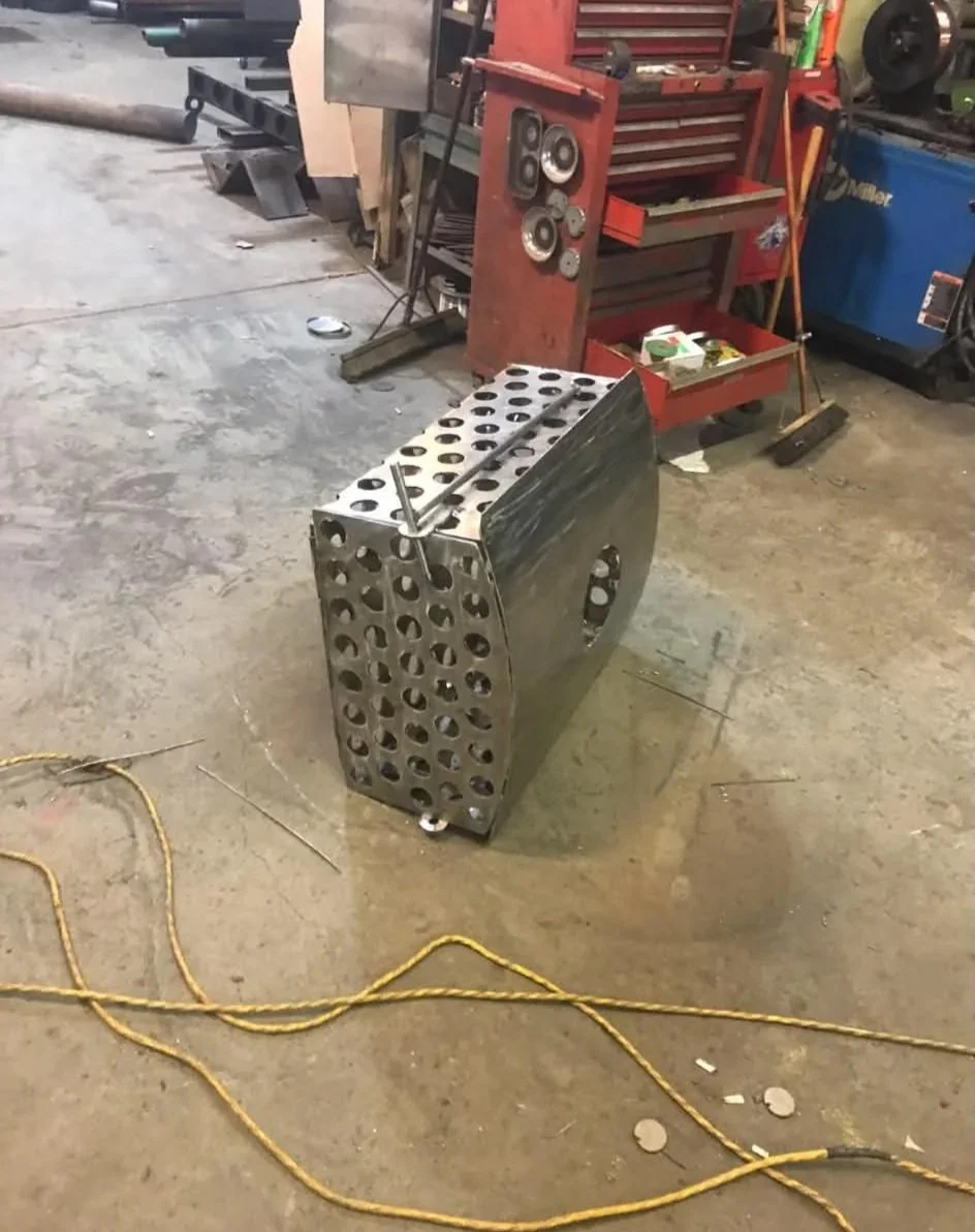 A metal work piece with multiple circular openings on a workshop floor, with tools and equipment in the background.