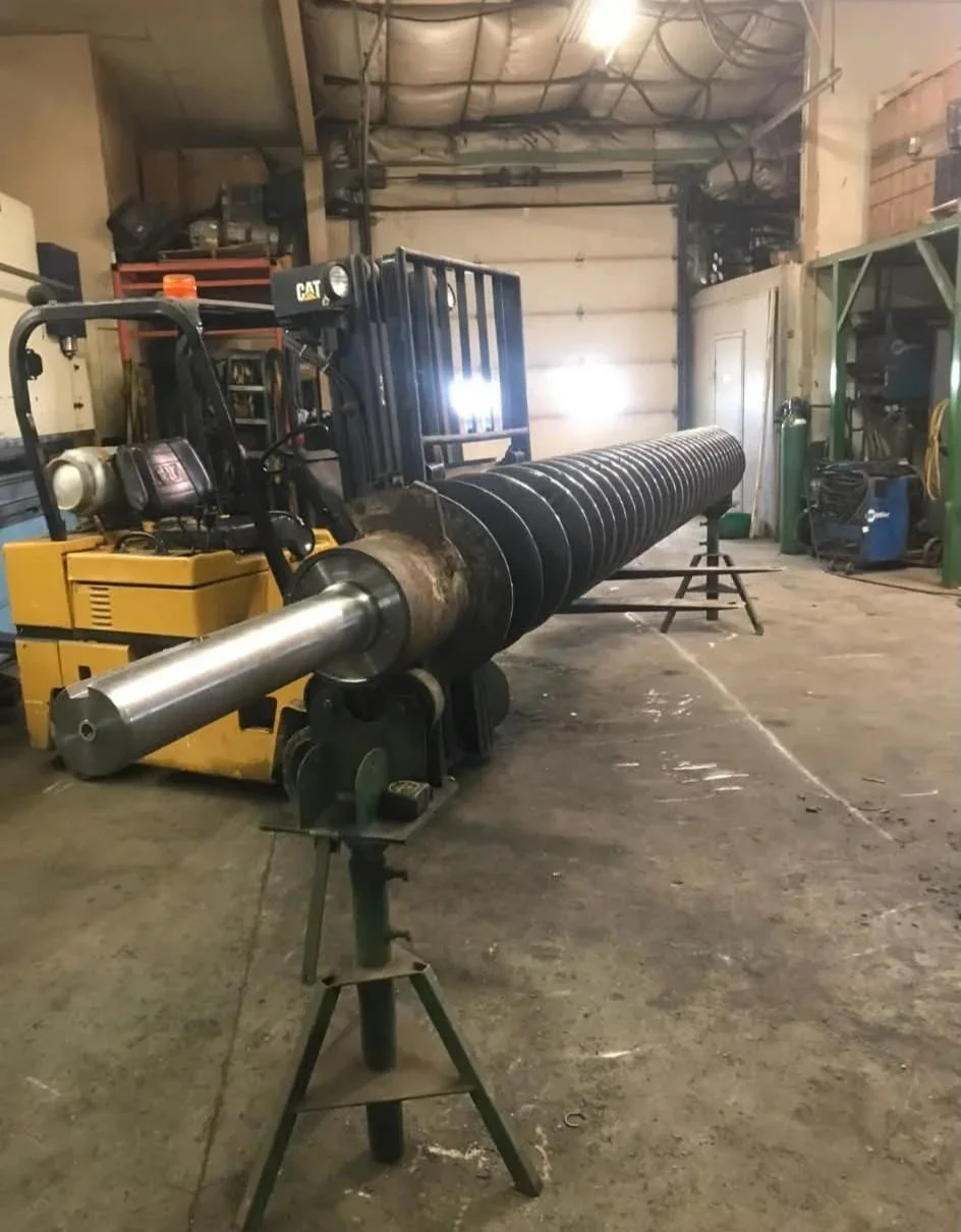 A large industrial drill pipe on a workbench in a workshop. The drill pipe extends into the background, with a yellow CAT machine nearby. The workshop has a concrete floor and walls, and various tools and equipment are visible in the background.