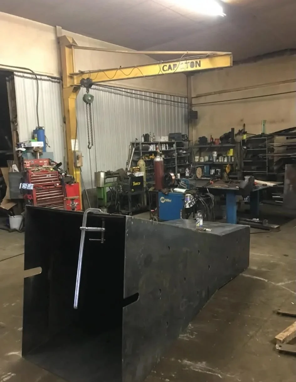A workshop with a yellow overhead crane labeled 'CAPE TOWN,' metal shelves, a workbench with tools, a welding machine, and a large black metal piece with a water fountain on top.