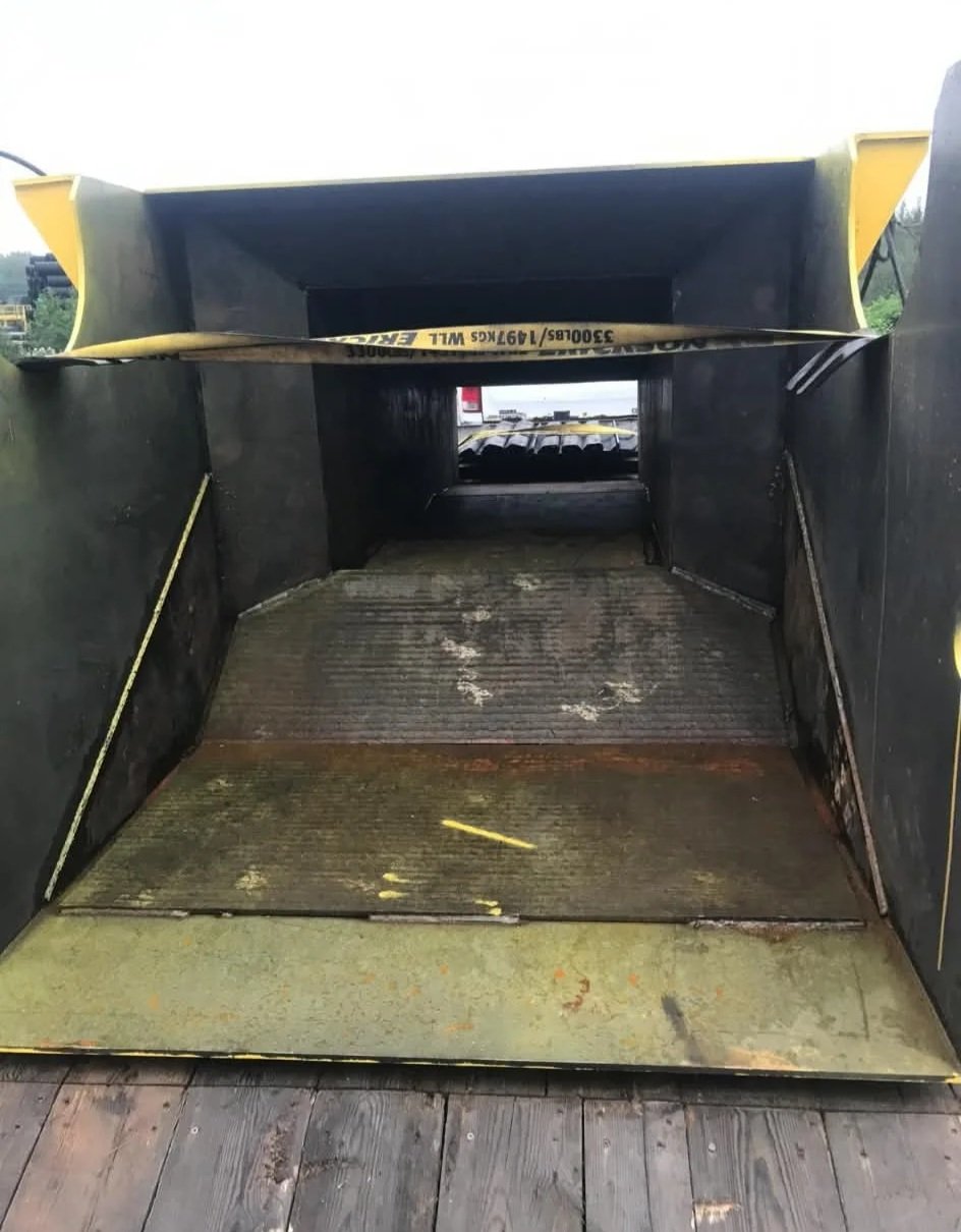 The photo shows the interior of a large industrial wheel loader bucket, with wear marks, dirt, and rust on the metal surface, and a yellow safety strap inside.
